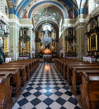 Zdjęcie nr 8: Kościół jest murowany z cegły, tynkowany, jednonawowy. Założony jest na rzucie prostokąta, z czteroprzęsłowym korpusem oraz jednoprzęsłowym prezbiterium, zamknięty prosto i skierowanym na południe. Ściana południowa wychodzi na ogród klasztorny, natomiast wschodnia i północna złączone są z klasztorem. Prezbiterium po obu stronach flankowane jest zakrystiami, nad którymi mieszczą się małe chóry. Chóry otwierają się do pojedynczymi arkadami do prezbiterium z drewnianymi balustradami. Do dwuprzęsłowej nawy od północy przylegają kolejne dwa przęsła korpusu, które stanowią kruchtę i kaplicę klasztoru, a nad nimi wzniesiony jest chór zakonny, oddzielony kratą. Nawa artykułowana jest za pomocą zdwojonych filarów, dźwigających wydatne belkowanie i tworzących półkoliście zamknięte wnęki arkadowe na ołtarze. Korpus kościoła sklepiony jest kolebkowo z lunetami na gurtach, natomiast w prezbiterium i kruchcie znajdują się sklepienia krzyżowe. Ściana arkady tęczowej zamknięta jest półkoliście. Wnętrze kościoła doświetlają umieszczone ponad belkowaniem okna w kształcie stojących prostokątów, zamknięte półkoliście. Do kościoła prowadzą drzwi wejściowe od północy przez kruchtę, do której wejście znajduje się od zachodu przez dziedziniec. Kolejne dwa wejścia prowadzą od strony klasztory przez kruchtę i zakrystię. Fasada północna kościoła wtopiona jest w bryłę klasztoru. Od strony dziedzińca, prowadzącego do głównego wejścia widoczna jest strona zachodnia kościoła, artykułowa zdwojonymi pilastrami. Portal główny flankowany jest kolumnami na tle pilastrów, które podtrzymują belkowanie i przerwany półkoliście zamknięty przyczółek. Podpory wsparte są na wysokich cokołach. Drzwi wejściowe flankuje półkoliście zamknięta arkada wsparta na parze filarów. Kościół nakrywa dach siodłowy, nad drugim przęsłem korpusu od strony północnej znajduje się wieżyczka na sygnaturkę ze zdwojoną latarnią. Przed kościołem znajduje się dziedziniec oddzielony od ulicy klasycystycznym murem z bramą.