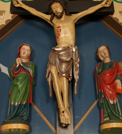 Zdjęcie nr 2: Grupa Ukrzyżowania złożona z figury Chrystusa przybitego do krzyża oraz dwóch rzeźb asystencyjnych, Matki Boskiej i św. Jana, ustawionych po jego dwóch stronach.
Chrystus ukazany jako umarły, przybity do krzyża trzema gwoździami. Ciało w zwisie, o rozłożonych ramionach; głowa przechylona na prawe ramię; sylwetka lekko esowato wygięta; prawa noga ugięta w kolanie, lewa wyprostowana; stopy w układzie prawa na lewą. Twarz pociągła, szczupła, o zamkniętych oczach, uniesionych brwiach, prostym nosie i wąskich ustach; okolona krótką, kręconą brodą oraz włosami skręconymi w pukle, opadającymi na ramiona. Ciało szczupłe, o zaznaczonej w sposób uproszczony muskulaturze i żebrach klatki piersiowej; ze śladami męki, w tym dużej rany w prawym boku, z której spływają plastyczne krople krwi. Na głowie korona cierniowa. Perizonium w postaci długiej tkaniny sięgającej kolan, ciężko opadające, modelowane z przodu w trzy duże fałdy w kształcie litery V. Po dwóch stronach bioder długie zwisy tkaniny układające się kaskadowo. Odsłonięte partie ciała polichromowane naturalistycznie, perizonium srebrzone. Krzyż o prostych zakończeniach ramion z titulusem w formie banderoli z napisem „INRI”.
Po lewej stronie krzyża figura Matki Boskiej ustawiona na wypukłej podstawie na planie owalu. Rzeźba ukazana frontalnie, o esowato wygiętej sylwetce, z głową przechyloną na lewe ramię, z ugiętą prawą nogą oraz uniesionymi, ugiętymi rękami na tle piersi, o splecionych dłoniach. Twarz ma pociągłą i szczupłą, o wysokim czole, migdałowatych oczach, prostym nosie i pełnych ustach, z podkreśloną brodą. Maria ubrana w długą, przylegającą, niebieską suknię, o łamanych fałdach skumulowanych u dołu i opadających obficie na podłoże, spośród których widoczne czubki spiczastych, czarnych butów. Na głowę i ramiona ma narzucony zielony płaszcz z czerwoną podszewką i srebrną lamówką, podtrzymywany rękami z przodu postaci; tkanina ściśle przylegająca do ciała, odzwierciedlająca układ sylwetki, silnie drapowana, układająca się z przodu w fałdy w kształcie litery V i proste opadające wzdłuż nóg, wokół głowy i na prawym boku brzegi płaszcza falowane, wokół prawej ręki tkanina wywinięta. Polichromia ciała naturalistyczna. 
Po prawej stronie krzyża figura św. Jana Ewangelisty ustawiona na wypukłej podstawie na planie owalu. Rzeźba ukazana frontalnie, o esowato wygiętej sylwetce, z głową przechyloną na prawe ramię, podtrzymywaną prawą ręką, druga ręką opuszczona wzdłuż ciała, w dłoni trzyma księgę. Twarz ma pociągłą i szczupłą, o wysokim czole, migdałowatych oczach, prostym nosie i pełnych ustach, z podkreśloną brodą; okoloną krótkimi włosami, kręconymi po bokach głowy, z grzywką nad czołem. Ubrany jest w długą, przylegającą, zieloną suknię o srebrnych lamówkach, obficie fałdowaną u dołu, opadającą na podłoże, z widocznymi bosymi stopami. Czerwony płaszcz o niebieskiej podszewce i szerokiej, srebrzonej lamówce zarzucony na ramiona i podtrzymywany prawą ręką z przodu postaci; obficie drapowany w fałdy o kształcie litery V z przodu, z wywiniętym brzegiem na piersi i na lewej ręce. Polichromia ciała naturalistyczna. 

