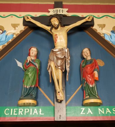 Zdjęcie nr 1: Grupa Ukrzyżowania złożona z figury Chrystusa przybitego do krzyża oraz dwóch rzeźb asystencyjnych, Matki Boskiej i św. Jana, ustawionych po jego dwóch stronach.
Chrystus ukazany jako umarły, przybity do krzyża trzema gwoździami. Ciało w zwisie, o rozłożonych ramionach; głowa przechylona na prawe ramię; sylwetka lekko esowato wygięta; prawa noga ugięta w kolanie, lewa wyprostowana; stopy w układzie prawa na lewą. Twarz pociągła, szczupła, o zamkniętych oczach, uniesionych brwiach, prostym nosie i wąskich ustach; okolona krótką, kręconą brodą oraz włosami skręconymi w pukle, opadającymi na ramiona. Ciało szczupłe, o zaznaczonej w sposób uproszczony muskulaturze i żebrach klatki piersiowej; ze śladami męki, w tym dużej rany w prawym boku, z której spływają plastyczne krople krwi. Na głowie korona cierniowa. Perizonium w postaci długiej tkaniny sięgającej kolan, ciężko opadające, modelowane z przodu w trzy duże fałdy w kształcie litery V. Po dwóch stronach bioder długie zwisy tkaniny układające się kaskadowo. Odsłonięte partie ciała polichromowane naturalistycznie, perizonium srebrzone. Krzyż o prostych zakończeniach ramion z titulusem w formie banderoli z napisem „INRI”.
Po lewej stronie krzyża figura Matki Boskiej ustawiona na wypukłej podstawie na planie owalu. Rzeźba ukazana frontalnie, o esowato wygiętej sylwetce, z głową przechyloną na lewe ramię, z ugiętą prawą nogą oraz uniesionymi, ugiętymi rękami na tle piersi, o splecionych dłoniach. Twarz ma pociągłą i szczupłą, o wysokim czole, migdałowatych oczach, prostym nosie i pełnych ustach, z podkreśloną brodą. Maria ubrana w długą, przylegającą, niebieską suknię, o łamanych fałdach skumulowanych u dołu i opadających obficie na podłoże, spośród których widoczne czubki spiczastych, czarnych butów. Na głowę i ramiona ma narzucony zielony płaszcz z czerwoną podszewką i srebrną lamówką, podtrzymywany rękami z przodu postaci; tkanina ściśle przylegająca do ciała, odzwierciedlająca układ sylwetki, silnie drapowana, układająca się z przodu w fałdy w kształcie litery V i proste opadające wzdłuż nóg, wokół głowy i na prawym boku brzegi płaszcza falowane, wokół prawej ręki tkanina wywinięta. Polichromia ciała naturalistyczna. 
Po prawej stronie krzyża figura św. Jana Ewangelisty ustawiona na wypukłej podstawie na planie owalu. Rzeźba ukazana frontalnie, o esowato wygiętej sylwetce, z głową przechyloną na prawe ramię, podtrzymywaną prawą ręką, druga ręką opuszczona wzdłuż ciała, w dłoni trzyma księgę. Twarz ma pociągłą i szczupłą, o wysokim czole, migdałowatych oczach, prostym nosie i pełnych ustach, z podkreśloną brodą; okoloną krótkimi włosami, kręconymi po bokach głowy, z grzywką nad czołem. Ubrany jest w długą, przylegającą, zieloną suknię o srebrnych lamówkach, obficie fałdowaną u dołu, opadającą na podłoże, z widocznymi bosymi stopami. Czerwony płaszcz o niebieskiej podszewce i szerokiej, srebrzonej lamówce zarzucony na ramiona i podtrzymywany prawą ręką z przodu postaci; obficie drapowany w fałdy o kształcie litery V z przodu, z wywiniętym brzegiem na piersi i na lewej ręce. Polichromia ciała naturalistyczna. 
