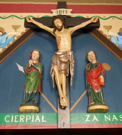 Zdjęcie nr 1: Grupa Ukrzyżowania złożona z figury Chrystusa przybitego do krzyża oraz dwóch rzeźb asystencyjnych, Matki Boskiej i św. Jana, ustawionych po jego dwóch stronach.
Chrystus ukazany jako umarły, przybity do krzyża trzema gwoździami. Ciało w zwisie, o rozłożonych ramionach; głowa przechylona na prawe ramię; sylwetka lekko esowato wygięta; prawa noga ugięta w kolanie, lewa wyprostowana; stopy w układzie prawa na lewą. Twarz pociągła, szczupła, o zamkniętych oczach, uniesionych brwiach, prostym nosie i wąskich ustach; okolona krótką, kręconą brodą oraz włosami skręconymi w pukle, opadającymi na ramiona. Ciało szczupłe, o zaznaczonej w sposób uproszczony muskulaturze i żebrach klatki piersiowej; ze śladami męki, w tym dużej rany w prawym boku, z której spływają plastyczne krople krwi. Na głowie korona cierniowa. Perizonium w postaci długiej tkaniny sięgającej kolan, ciężko opadające, modelowane z przodu w trzy duże fałdy w kształcie litery V. Po dwóch stronach bioder długie zwisy tkaniny układające się kaskadowo. Odsłonięte partie ciała polichromowane naturalistycznie, perizonium srebrzone. Krzyż o prostych zakończeniach ramion z titulusem w formie banderoli z napisem „INRI”.
Po lewej stronie krzyża figura Matki Boskiej ustawiona na wypukłej podstawie na planie owalu. Rzeźba ukazana frontalnie, o esowato wygiętej sylwetce, z głową przechyloną na lewe ramię, z ugiętą prawą nogą oraz uniesionymi, ugiętymi rękami na tle piersi, o splecionych dłoniach. Twarz ma pociągłą i szczupłą, o wysokim czole, migdałowatych oczach, prostym nosie i pełnych ustach, z podkreśloną brodą. Maria ubrana w długą, przylegającą, niebieską suknię, o łamanych fałdach skumulowanych u dołu i opadających obficie na podłoże, spośród których widoczne czubki spiczastych, czarnych butów. Na głowę i ramiona ma narzucony zielony płaszcz z czerwoną podszewką i srebrną lamówką, podtrzymywany rękami z przodu postaci; tkanina ściśle przylegająca do ciała, odzwierciedlająca układ sylwetki, silnie drapowana, układająca się z przodu w fałdy w kształcie litery V i proste opadające wzdłuż nóg, wokół głowy i na prawym boku brzegi płaszcza falowane, wokół prawej ręki tkanina wywinięta. Polichromia ciała naturalistyczna. 
Po prawej stronie krzyża figura św. Jana Ewangelisty ustawiona na wypukłej podstawie na planie owalu. Rzeźba ukazana frontalnie, o esowato wygiętej sylwetce, z głową przechyloną na prawe ramię, podtrzymywaną prawą ręką, druga ręką opuszczona wzdłuż ciała, w dłoni trzyma księgę. Twarz ma pociągłą i szczupłą, o wysokim czole, migdałowatych oczach, prostym nosie i pełnych ustach, z podkreśloną brodą; okoloną krótkimi włosami, kręconymi po bokach głowy, z grzywką nad czołem. Ubrany jest w długą, przylegającą, zieloną suknię o srebrnych lamówkach, obficie fałdowaną u dołu, opadającą na podłoże, z widocznymi bosymi stopami. Czerwony płaszcz o niebieskiej podszewce i szerokiej, srebrzonej lamówce zarzucony na ramiona i podtrzymywany prawą ręką z przodu postaci; obficie drapowany w fałdy o kształcie litery V z przodu, z wywiniętym brzegiem na piersi i na lewej ręce. Polichromia ciała naturalistyczna. 

