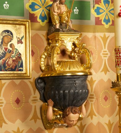 Zdjęcie nr 1: Półkolista misa o płaszczu z imitacją puklowania i brzegiem w formie profilowanego gzymsu. Ujęta uszami w kształcie wolut przytwierdzonych do ściany. Podstawę misy tworzy rzeźbiona półpostać anioła wyłaniającego się z obłoku, który dźwiga ją na plecach i ujmuje uniesionymi w górę rękami. Twarz ma pociągłą, o pełnych policzkach i wysokim czole, dużych, migdałowatych oczach, szerokim nosie i rozchylonych, wąskich ustach. Głowę okalają krótkie, silnie kręcone włosy. Anioł ubrany w złoconą tunikę z podwiniętymi rękawami. Skrzydła złocone, złożone. Pokrywa złożona z podstawy na planie ściętego koła, z wybrzuszeniem dekorowanym liśćmi, na niej cztery rocaille'owe kabłąki, dźwigające prostokątną podstawę, na której umieszczona jest niewielka grupa rzeźbiarska ze sceną Chrztu Chrystusa. Misa polichromowana na sposób imitujący marmoryzację. 

