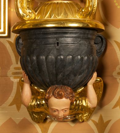 Zdjęcie nr 4: Półkolista misa o płaszczu z imitacją puklowania i brzegiem w formie profilowanego gzymsu. Ujęta uszami w kształcie wolut przytwierdzonych do ściany. Podstawę misy tworzy rzeźbiona półpostać anioła wyłaniającego się z obłoku, który dźwiga ją na plecach i ujmuje uniesionymi w górę rękami. Twarz ma pociągłą, o pełnych policzkach i wysokim czole, dużych, migdałowatych oczach, szerokim nosie i rozchylonych, wąskich ustach. Głowę okalają krótkie, silnie kręcone włosy. Anioł ubrany w złoconą tunikę z podwiniętymi rękawami. Skrzydła złocone, złożone. Pokrywa złożona z podstawy na planie ściętego koła, z wybrzuszeniem dekorowanym liśćmi, na niej cztery rocaille'owe kabłąki, dźwigające prostokątną podstawę, na której umieszczona jest niewielka grupa rzeźbiarska ze sceną Chrztu Chrystusa. Misa polichromowana na sposób imitujący marmoryzację. 

