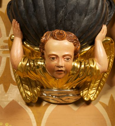 Zdjęcie nr 5: Półkolista misa o płaszczu z imitacją puklowania i brzegiem w formie profilowanego gzymsu. Ujęta uszami w kształcie wolut przytwierdzonych do ściany. Podstawę misy tworzy rzeźbiona półpostać anioła wyłaniającego się z obłoku, który dźwiga ją na plecach i ujmuje uniesionymi w górę rękami. Twarz ma pociągłą, o pełnych policzkach i wysokim czole, dużych, migdałowatych oczach, szerokim nosie i rozchylonych, wąskich ustach. Głowę okalają krótkie, silnie kręcone włosy. Anioł ubrany w złoconą tunikę z podwiniętymi rękawami. Skrzydła złocone, złożone. Pokrywa złożona z podstawy na planie ściętego koła, z wybrzuszeniem dekorowanym liśćmi, na niej cztery rocaille'owe kabłąki, dźwigające prostokątną podstawę, na której umieszczona jest niewielka grupa rzeźbiarska ze sceną Chrztu Chrystusa. Misa polichromowana na sposób imitujący marmoryzację. 

