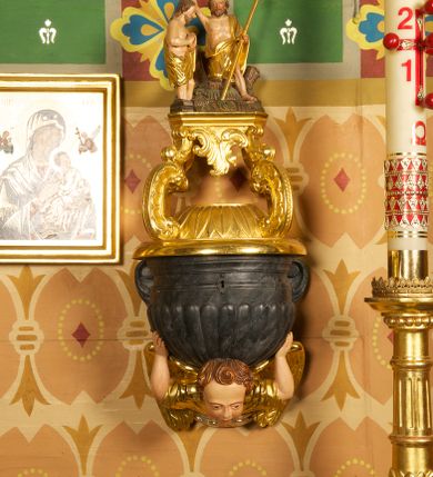 Zdjęcie nr 2: Półkolista misa o płaszczu z imitacją puklowania i brzegiem w formie profilowanego gzymsu. Ujęta uszami w kształcie wolut przytwierdzonych do ściany. Podstawę misy tworzy rzeźbiona półpostać anioła wyłaniającego się z obłoku, który dźwiga ją na plecach i ujmuje uniesionymi w górę rękami. Twarz ma pociągłą, o pełnych policzkach i wysokim czole, dużych, migdałowatych oczach, szerokim nosie i rozchylonych, wąskich ustach. Głowę okalają krótkie, silnie kręcone włosy. Anioł ubrany w złoconą tunikę z podwiniętymi rękawami. Skrzydła złocone, złożone. Pokrywa złożona z podstawy na planie ściętego koła, z wybrzuszeniem dekorowanym liśćmi, na niej cztery rocaille'owe kabłąki, dźwigające prostokątną podstawę, na której umieszczona jest niewielka grupa rzeźbiarska ze sceną Chrztu Chrystusa. Misa polichromowana na sposób imitujący marmoryzację. 

