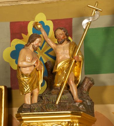 Zdjęcie nr 6: Półkolista misa o płaszczu z imitacją puklowania i brzegiem w formie profilowanego gzymsu. Ujęta uszami w kształcie wolut przytwierdzonych do ściany. Podstawę misy tworzy rzeźbiona półpostać anioła wyłaniającego się z obłoku, który dźwiga ją na plecach i ujmuje uniesionymi w górę rękami. Twarz ma pociągłą, o pełnych policzkach i wysokim czole, dużych, migdałowatych oczach, szerokim nosie i rozchylonych, wąskich ustach. Głowę okalają krótkie, silnie kręcone włosy. Anioł ubrany w złoconą tunikę z podwiniętymi rękawami. Skrzydła złocone, złożone. Pokrywa złożona z podstawy na planie ściętego koła, z wybrzuszeniem dekorowanym liśćmi, na niej cztery rocaille'owe kabłąki, dźwigające prostokątną podstawę, na której umieszczona jest niewielka grupa rzeźbiarska ze sceną Chrztu Chrystusa. Misa polichromowana na sposób imitujący marmoryzację. 

