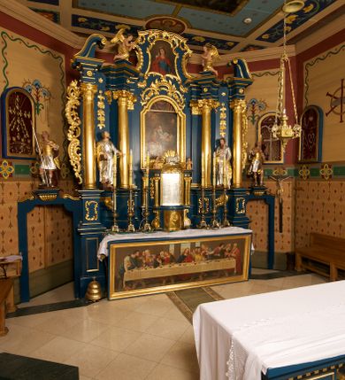 Zdjęcie nr 2: Ołtarz drewniany, prostopadłościenny, z kamienną mensą. Antepedium w formie obrazu z przedstawieniem Ostatniej Wieczerzy, ujętego profilowaną ramą. Na ołtarzu ustawione tabernakulum z tronem wystawienia. 
Nastawa architektoniczna, jednokondygnacyjna ze zwieńczeniem, trójosiowa z bramkami, ustawiona na dwustrefowym cokole z wyłamanymi czterema prostopadłościanami. Oś środkowa ujęta dwiema parami pilastrów oraz parą kolumn, na cokołach zwróconych pod kątem do wewnątrz ołtarza. Osie zewnętrzne ujęte parą pilastrów i stojących na ich tle kolumn. Wszystkie podpory dźwigają imposty, na osiach środkowy podpór zwrócone pod kątem do wewnątrz. Na parze skrajnych impostów fragmenty przerwanego, falistego przyczółka. W polu środkowym nisza w formie stojącego prostokąta zamkniętego łukiem półkolistym nadwieszonym, z uskokiem, ujęta profilowaną ramą, z obrazem Nauczania Marii. Na osiach bocznych figury św. Piotra i św. Pawła ustawione na wspornikach Zwieńczenie niearchitektoniczne, o formie zbliżonej do trapezu, o wklęsłych bokach z uszakami, zamknięte fragmentem falistego gzymsu. W polu obraz o fantazyjnej formie zbliżonej do trapezu z przedstawieniem św. Barbary, ujęty ornamentalną ramą. Zwieńczenie flankują dwie figury klęczących aniołów na cokołach. Nastawa dekorowana rocaille&#039;m w przestrzeni nad polem głównym oraz w partii zwieńczenia, ujęta uszami utworzonymi z rocaille&#039;u, kampanuli i ornamentu roślinnego. Na osiach bocznych zwisy kwiatowe. Bramki w formie przejść o wykroju stojącego prostokąta o wklęsłych górnych narożach, zamkniętego dwiema woltami i cokołami z imitacją puklowania, na których stoją figury niezidentyfikowanych monarchów. Struktura polichromowana w kolorze niebieskim, kolumny, profile i ornamentyka złocone. 






