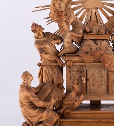 Zdjęcie nr 5: Tabernakulum w formie Arki Przymierza, adorowanej przez dwie klęczące figury św. Jana i starotestamentowego arcykapłana, dwie stojące postacie lewitów oraz klęczących aniołów, zwieńczonej glorią promienistą z okiem Opatrzności. Arka w formie prostopadłościennej szafki z dwoma drążkami do noszenia jest od frontu podzielone na trzy pola: środkowe zamknięte łukiem wklęsło-wypukłym i boczne o ćwierćkoliście wyciętych narożach. Pole środkowe stanowią drzwiczki z rytymi cyframi Dekalogu, a w bocznych znajdują się kłosy zbóż i winne grono. Na skłębionych chmurach z podestem leży baranek. Nad nim widnieje znacznej wielkości gloria promienista z wieńcem chmur i umieszczonym w nim okiem Opatrzności oraz cztery uskrzydlone główki anielskie. Nad parą główek w górnej części papieska tiara ze skrzyżowanymi kluczami. Po bokach baranka dwa klęczące, uskrzydlone nagie aniołki o rękach i nogach utworzonych z piór. Obok arki dwie postacie lewitów ubranych w długie szaty, w różnicowanych pozach i gestach. Poniżej klęczą postacie św. Jana z księgą i orłem obok niego i kapłana w tradycyjnym starotestamentowym stroju – efodzie czyli tunice i płaszczu wierzchnim przepasanym na biodrach, z pektorałem na piersi i czapką na głowie. 