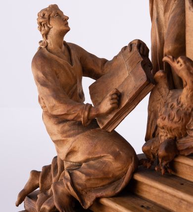 Zdjęcie nr 15: Tabernakulum w formie Arki Przymierza, adorowanej przez dwie klęczące figury św. Jana i starotestamentowego arcykapłana, dwie stojące postacie lewitów oraz klęczących aniołów, zwieńczonej glorią promienistą z okiem Opatrzności. Arka w formie prostopadłościennej szafki z dwoma drążkami do noszenia jest od frontu podzielone na trzy pola: środkowe zamknięte łukiem wklęsło-wypukłym i boczne o ćwierćkoliście wyciętych narożach. Pole środkowe stanowią drzwiczki z rytymi cyframi Dekalogu, a w bocznych znajdują się kłosy zbóż i winne grono. Na skłębionych chmurach z podestem leży baranek. Nad nim widnieje znacznej wielkości gloria promienista z wieńcem chmur i umieszczonym w nim okiem Opatrzności oraz cztery uskrzydlone główki anielskie. Nad parą główek w górnej części papieska tiara ze skrzyżowanymi kluczami. Po bokach baranka dwa klęczące, uskrzydlone nagie aniołki o rękach i nogach utworzonych z piór. Obok arki dwie postacie lewitów ubranych w długie szaty, w różnicowanych pozach i gestach. Poniżej klęczą postacie św. Jana z księgą i orłem obok niego i kapłana w tradycyjnym starotestamentowym stroju – efodzie czyli tunice i płaszczu wierzchnim przepasanym na biodrach, z pektorałem na piersi i czapką na głowie. 