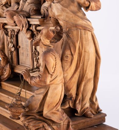 Zdjęcie nr 18: Tabernakulum w formie Arki Przymierza, adorowanej przez dwie klęczące figury św. Jana i starotestamentowego arcykapłana, dwie stojące postacie lewitów oraz klęczących aniołów, zwieńczonej glorią promienistą z okiem Opatrzności. Arka w formie prostopadłościennej szafki z dwoma drążkami do noszenia jest od frontu podzielone na trzy pola: środkowe zamknięte łukiem wklęsło-wypukłym i boczne o ćwierćkoliście wyciętych narożach. Pole środkowe stanowią drzwiczki z rytymi cyframi Dekalogu, a w bocznych znajdują się kłosy zbóż i winne grono. Na skłębionych chmurach z podestem leży baranek. Nad nim widnieje znacznej wielkości gloria promienista z wieńcem chmur i umieszczonym w nim okiem Opatrzności oraz cztery uskrzydlone główki anielskie. Nad parą główek w górnej części papieska tiara ze skrzyżowanymi kluczami. Po bokach baranka dwa klęczące, uskrzydlone nagie aniołki o rękach i nogach utworzonych z piór. Obok arki dwie postacie lewitów ubranych w długie szaty, w różnicowanych pozach i gestach. Poniżej klęczą postacie św. Jana z księgą i orłem obok niego i kapłana w tradycyjnym starotestamentowym stroju – efodzie czyli tunice i płaszczu wierzchnim przepasanym na biodrach, z pektorałem na piersi i czapką na głowie. 