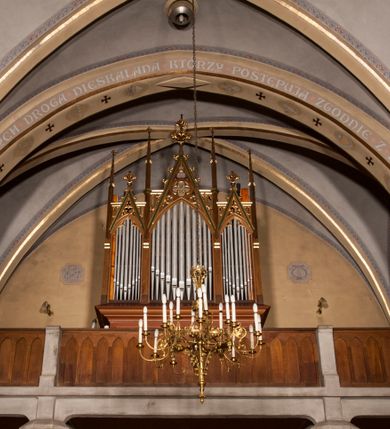 Zdjęcie nr 15: Kościół ceglany z kamiennym przyziemiem, na planie prostokąta z węższym i niższym prezbiterium flankowanym dwoma aneksami wystającymi poza lico nawy i wieżą od zachodu. Kościół orientowany, jednonawowy, czteroprzęsłowy z węższym, jednoprzęsłowym prezbiterium zamkniętym trójbocznie, z kruchtą w przyziemiu wieży. Nawa artykułowana półfilarami o ściętych narożach, przechodzących w gurty krzyżowo-żebrowego sklepienia. W nawie i prezbiterium okna prostokątne zamknięte łukiem ostrym; główne wejście od wschodu. Identyczne okna w aneksach przy prezbiterium. Aneksy dwuprzęsłowe ujęte wielobocznymi filarami, zamknięte trójbocznie z parasolowym sklepieniem. Chór muzyczny w czwartym przęśle nawy, wsparty na wielobocznych filarach z drewnianym parapetem ozdobionym ostrołukowymi blendami. Kruchta węższa od nawy, zasklepiona krzyżowo. Fasada węższa od nawy, ujęta przyporami w narożach, między którymi ostrołukowe wejście. W drugiej kondygnacji oddzielonej parapetem wysokie okno zamknięte łukiem ostrym, nad którym tarcza zegara. Nad fasadą ośmioboczna wieża. W wieży na przemian okna i płyciny, zamknięte łukiem ostrym. Trójboczne prezbiterium w narożach ujęte przyporami sięgającymi połowy wysokości, między którymi ostrołukowe okna. Aneksy na planie prostokąta z przyporami na narożach oraz jedną w części bocznej. Czteroosiowa ściana nawy i naroża oddzielone przyporami, między którymi ostrołukowo zakończone okna. Hełm wieży w formie ośmiobocznego stożka, załamującego się w partii dolnej. Nawa z dwuspadowym dachem z wieżyczką na sygnaturkę nad pierwszym przęsłem.