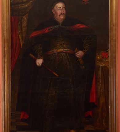 Zdjęcie nr 1: Portret w kształcie stojącego prostokąta z przedstawieniem króla Jana III Sobieskiego w centrum kompozycji. Król ukazany w postawie stojącej z prawą nogą wysuniętą do przodu, w prawej dłoni trzyma regiment, a lewą wspiera na rękojeści szabli, przypiętej do pasa. Twarz owalna z długim nosem i małymi oczami, z wąsami i wyraźnie zaznaczonym podbródkiem. Sobieski ubrany jest w oliwkowozielony żupan, przewiązany pasem kontuszowym oraz czerwony płaszcz – delię – z ciemną podszewką oraz żółte buty. Tło ciemne, u góry i po lewej stronie obrazu podwieszona jest kotara, u doły podłoga w kratownicę, a po prawej stronie stoi stolik, nakryty złotym obrusem. Na stoliku leżą insygnia królewskie: berło, jabłko i korona. Wyżej czerwona tarcza herbowa z koroną i godłem Królestwa – Orłem Białym z tarczą na piersi. Rama drewniana, profilowana, złocona. Na odwrociu karteczka z drukowanym napisem „Ze zbiorów księdza Jana kniazia z Kozielska Puzyny księcia biskupa krakowskiego, no 45”.

