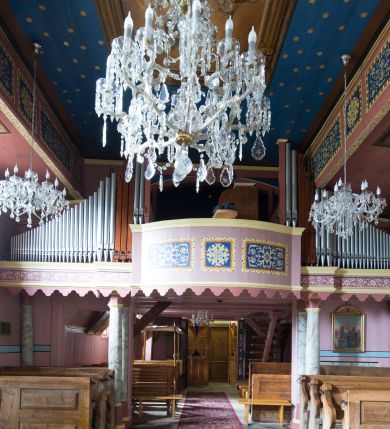 Zdjęcie nr 7: Kościół jest orientowany, drewniany, wzniesiony w konstrukcji zrębowej, o ścianach oszalowanych pionowo, z listwowaniem. Do korpusu nawowego, na rzucie zbliżonym do kwadratu, od wschodu przylega węższe, kwadratowe prezbiterium z dwukondygnacyjną zakrystią ze skarbcem od północy. Od zachodu do korpusu dobudowana jest wieża o konstrukcji słupowo-ramowej i pochyłych ścianach, z przedsionkiem w przyziemiu oraz z pozorną izbicą. Otoczona jest szalowanymi sobotami. Do wieży od zachodu i do korpusu od południa przylegają kruchty. Wnętrze przekryte jest stropami, w nawie z zaskrzynieniami. 
Korpus nawowy, prezbiterium i kruchty nakryte dachami dwuspadowymi, soboty pulpitowymi. Wszystkie dachy kryte gontem. Wieżę wieńczy baniasty hełm, kalenicę nad korpusem sygnaturka z latarnią. 
Ściany wnętrza zdobi figuralno-ornamentalna polichromia. Po zachodniej stronie korpusu nawowego usytuowany jest chór muzyczny wsparty na dwóch kolumnach.
Plac przykościelny otoczony jest murowanym ogrodzeniem oraz wieńcem starych lip.  
