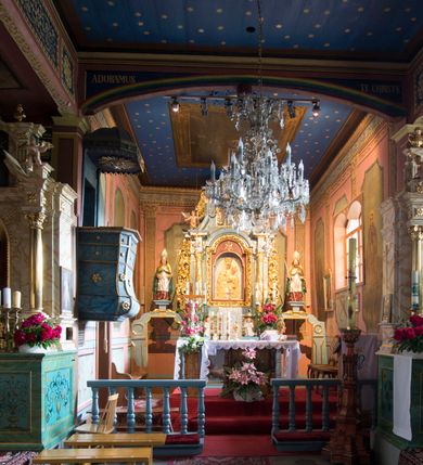 Zdjęcie nr 10: Kościół jest orientowany, drewniany, wzniesiony w konstrukcji zrębowej, o ścianach oszalowanych pionowo, z listwowaniem. Do korpusu nawowego, na rzucie zbliżonym do kwadratu, od wschodu przylega węższe, kwadratowe prezbiterium z dwukondygnacyjną zakrystią ze skarbcem od północy. Od zachodu do korpusu dobudowana jest wieża o konstrukcji słupowo-ramowej i pochyłych ścianach, z przedsionkiem w przyziemiu oraz z pozorną izbicą. Otoczona jest szalowanymi sobotami. Do wieży od zachodu i do korpusu od południa przylegają kruchty. Wnętrze przekryte jest stropami, w nawie z zaskrzynieniami. 
Korpus nawowy, prezbiterium i kruchty nakryte dachami dwuspadowymi, soboty pulpitowymi. Wszystkie dachy kryte gontem. Wieżę wieńczy baniasty hełm, kalenicę nad korpusem sygnaturka z latarnią. 
Ściany wnętrza zdobi figuralno-ornamentalna polichromia. Po zachodniej stronie korpusu nawowego usytuowany jest chór muzyczny wsparty na dwóch kolumnach.
Plac przykościelny otoczony jest murowanym ogrodzeniem oraz wieńcem starych lip.  
