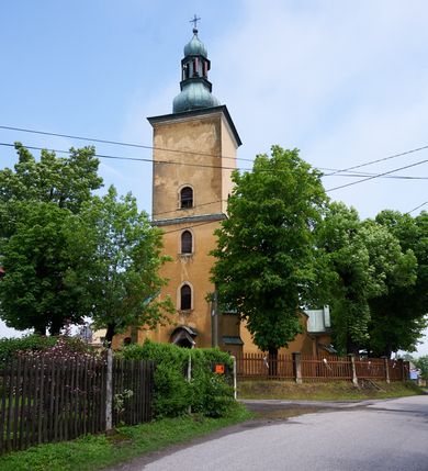 Zdjęcie nr 2: Kościół murowany z cegły i kamienia, tynkowany. Zlokalizowany po północnej stronie wiejskiej drogi, na placu wydzielonym drewnianym płotem rozpiętym pomiędzy murowanymi, tynkowanymi filarami. Budowla orientowana, wzniesiona na wydłużonym rzucie złożonym z krótkiego, zamkniętego wielobocznie prezbiterium, nawy na rzucie zbliżonym  do kwadratu z centralnie umieszczonym filarem i wieży na rzucie kwadratu. Od północy do prezbiterium przylega zakrystia na rzucie kwadratu, do nawy kaplica na rzucie prostokąta. Do wieży dostawiony aneks z klatką schodową prowadzącą na chór. Od południa do nawy przylega kruchta na rzucie kwadratu. Całość budowli oszkarpowana w narożach. Wnętrze nakrywają sklepienia krzyżowe, w nawie żebra wsparte są na czworobocznym, murowanym filarze centralnym. W kruchcie pod wieżą sklepienie kryształowe, w zakrystii sieciowe. Otwory drzwiowe o wykroju ostrołukowym, w kamiennych portalach. Okna o wykroju stojących prostokątów, zwieńczonych łukiem pełnym; w północnej ścianie kaplicy okulus. Elewacje świątyni opracowane jednolicie, tynkowane, z niezbyt wydatnym gzymsem podokapowym, w narożach opięte masywnymi, trójuskokowymi przyporami. Elewacja zachodnia, wieżowa, czterokondygnacyjna, jednoosiowa, flankowana parą przypór. Gzyms kordonowy wydziela ostatnią, najwyższa kondygnację. W osi portal kamienny, zamknięty łukiem półkolistym. Powyżej, w osi okna w każdej z kondygnacji. Na pierwszym piętrze, w zworniku obramienia okiennego herb Drużyna w kartuszu. Boczne elewacje wieży bez otworów okiennych. Elewacja północna artykułowana przyporami, o nieregularnym układzie osi, przesłonięta przybudówkami kaplicy, zakrystii i klatki schodowej wiodącej na chór. W elewacji kaplicy, w osi okulus. W części prezbiterialnej okna o wykroju prostokąta zamkniętego łukiem półkolistym w dekoracyjnych obramieniach kamiennych z pilasterkami i archiwoltą zdobione płycinami i dekoracją ornamentalną w formie rozet i herbem Drużyna. Elewacja wschodnia artykułowana przyporami, przesłonięta parawanową ścianką drewnianą o trzech przeszklonych oknach-arkadah; w głębi ogrójec. Elewacja południowa artykułowana przyporami. W osi korpusu nawowego kruchta, ponad nią, po bokach dwa okna z obramieniami kamiennymi, dekorowanymi ornamentalnie i herbem Drużyna.  Dachy dwuspadowe, kryte blachą. Nieco wyższy nad korpusem nawowym, niższy nad prezbiterium. Nad zakrystią dach pulpitowy. Wieża zwieńczona cebulastym hełmem z latarnią.