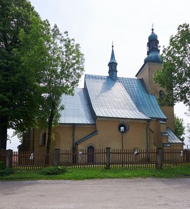 Zdjęcie nr 15: Kościół murowany z cegły i kamienia, tynkowany. Zlokalizowany po północnej stronie wiejskiej drogi, na placu wydzielonym drewnianym płotem rozpiętym pomiędzy murowanymi, tynkowanymi filarami. Budowla orientowana, wzniesiona na wydłużonym rzucie złożonym z krótkiego, zamkniętego wielobocznie prezbiterium, nawy na rzucie zbliżonym  do kwadratu z centralnie umieszczonym filarem i wieży na rzucie kwadratu. Od północy do prezbiterium przylega zakrystia na rzucie kwadratu, do nawy kaplica na rzucie prostokąta. Do wieży dostawiony aneks z klatką schodową prowadzącą na chór. Od południa do nawy przylega kruchta na rzucie kwadratu. Całość budowli oszkarpowana w narożach. Wnętrze nakrywają sklepienia krzyżowe, w nawie żebra wsparte są na czworobocznym, murowanym filarze centralnym. W kruchcie pod wieżą sklepienie kryształowe, w zakrystii sieciowe. Otwory drzwiowe o wykroju ostrołukowym, w kamiennych portalach. Okna o wykroju stojących prostokątów, zwieńczonych łukiem pełnym; w północnej ścianie kaplicy okulus. Elewacje świątyni opracowane jednolicie, tynkowane, z niezbyt wydatnym gzymsem podokapowym, w narożach opięte masywnymi, trójuskokowymi przyporami. Elewacja zachodnia, wieżowa, czterokondygnacyjna, jednoosiowa, flankowana parą przypór. Gzyms kordonowy wydziela ostatnią, najwyższa kondygnację. W osi portal kamienny, zamknięty łukiem półkolistym. Powyżej, w osi okna w każdej z kondygnacji. Na pierwszym piętrze, w zworniku obramienia okiennego herb Drużyna w kartuszu. Boczne elewacje wieży bez otworów okiennych. Elewacja północna artykułowana przyporami, o nieregularnym układzie osi, przesłonięta przybudówkami kaplicy, zakrystii i klatki schodowej wiodącej na chór. W elewacji kaplicy, w osi okulus. W części prezbiterialnej okna o wykroju prostokąta zamkniętego łukiem półkolistym w dekoracyjnych obramieniach kamiennych z pilasterkami i archiwoltą zdobione płycinami i dekoracją ornamentalną w formie rozet i herbem Drużyna. Elewacja wschodnia artykułowana przyporami, przesłonięta parawanową ścianką drewnianą o trzech przeszklonych oknach-arkadah; w głębi ogrójec. Elewacja południowa artykułowana przyporami. W osi korpusu nawowego kruchta, ponad nią, po bokach dwa okna z obramieniami kamiennymi, dekorowanymi ornamentalnie i herbem Drużyna.  Dachy dwuspadowe, kryte blachą. Nieco wyższy nad korpusem nawowym, niższy nad prezbiterium. Nad zakrystią dach pulpitowy. Wieża zwieńczona cebulastym hełmem z latarnią.