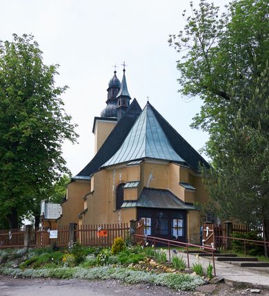 Zdjęcie nr 16: Kościół murowany z cegły i kamienia, tynkowany. Zlokalizowany po północnej stronie wiejskiej drogi, na placu wydzielonym drewnianym płotem rozpiętym pomiędzy murowanymi, tynkowanymi filarami. Budowla orientowana, wzniesiona na wydłużonym rzucie złożonym z krótkiego, zamkniętego wielobocznie prezbiterium, nawy na rzucie zbliżonym  do kwadratu z centralnie umieszczonym filarem i wieży na rzucie kwadratu. Od północy do prezbiterium przylega zakrystia na rzucie kwadratu, do nawy kaplica na rzucie prostokąta. Do wieży dostawiony aneks z klatką schodową prowadzącą na chór. Od południa do nawy przylega kruchta na rzucie kwadratu. Całość budowli oszkarpowana w narożach. Wnętrze nakrywają sklepienia krzyżowe, w nawie żebra wsparte są na czworobocznym, murowanym filarze centralnym. W kruchcie pod wieżą sklepienie kryształowe, w zakrystii sieciowe. Otwory drzwiowe o wykroju ostrołukowym, w kamiennych portalach. Okna o wykroju stojących prostokątów, zwieńczonych łukiem pełnym; w północnej ścianie kaplicy okulus. Elewacje świątyni opracowane jednolicie, tynkowane, z niezbyt wydatnym gzymsem podokapowym, w narożach opięte masywnymi, trójuskokowymi przyporami. Elewacja zachodnia, wieżowa, czterokondygnacyjna, jednoosiowa, flankowana parą przypór. Gzyms kordonowy wydziela ostatnią, najwyższa kondygnację. W osi portal kamienny, zamknięty łukiem półkolistym. Powyżej, w osi okna w każdej z kondygnacji. Na pierwszym piętrze, w zworniku obramienia okiennego herb Drużyna w kartuszu. Boczne elewacje wieży bez otworów okiennych. Elewacja północna artykułowana przyporami, o nieregularnym układzie osi, przesłonięta przybudówkami kaplicy, zakrystii i klatki schodowej wiodącej na chór. W elewacji kaplicy, w osi okulus. W części prezbiterialnej okna o wykroju prostokąta zamkniętego łukiem półkolistym w dekoracyjnych obramieniach kamiennych z pilasterkami i archiwoltą zdobione płycinami i dekoracją ornamentalną w formie rozet i herbem Drużyna. Elewacja wschodnia artykułowana przyporami, przesłonięta parawanową ścianką drewnianą o trzech przeszklonych oknach-arkadah; w głębi ogrójec. Elewacja południowa artykułowana przyporami. W osi korpusu nawowego kruchta, ponad nią, po bokach dwa okna z obramieniami kamiennymi, dekorowanymi ornamentalnie i herbem Drużyna.  Dachy dwuspadowe, kryte blachą. Nieco wyższy nad korpusem nawowym, niższy nad prezbiterium. Nad zakrystią dach pulpitowy. Wieża zwieńczona cebulastym hełmem z latarnią.