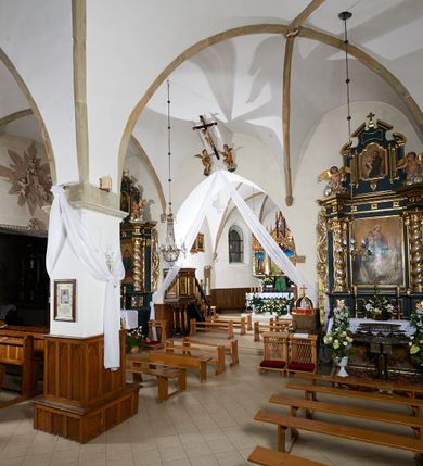 Zdjęcie nr 3: Kościół murowany z cegły i kamienia, tynkowany. Zlokalizowany po północnej stronie wiejskiej drogi, na placu wydzielonym drewnianym płotem rozpiętym pomiędzy murowanymi, tynkowanymi filarami. Budowla orientowana, wzniesiona na wydłużonym rzucie złożonym z krótkiego, zamkniętego wielobocznie prezbiterium, nawy na rzucie zbliżonym  do kwadratu z centralnie umieszczonym filarem i wieży na rzucie kwadratu. Od północy do prezbiterium przylega zakrystia na rzucie kwadratu, do nawy kaplica na rzucie prostokąta. Do wieży dostawiony aneks z klatką schodową prowadzącą na chór. Od południa do nawy przylega kruchta na rzucie kwadratu. Całość budowli oszkarpowana w narożach. Wnętrze nakrywają sklepienia krzyżowe, w nawie żebra wsparte są na czworobocznym, murowanym filarze centralnym. W kruchcie pod wieżą sklepienie kryształowe, w zakrystii sieciowe. Otwory drzwiowe o wykroju ostrołukowym, w kamiennych portalach. Okna o wykroju stojących prostokątów, zwieńczonych łukiem pełnym; w północnej ścianie kaplicy okulus. Elewacje świątyni opracowane jednolicie, tynkowane, z niezbyt wydatnym gzymsem podokapowym, w narożach opięte masywnymi, trójuskokowymi przyporami. Elewacja zachodnia, wieżowa, czterokondygnacyjna, jednoosiowa, flankowana parą przypór. Gzyms kordonowy wydziela ostatnią, najwyższa kondygnację. W osi portal kamienny, zamknięty łukiem półkolistym. Powyżej, w osi okna w każdej z kondygnacji. Na pierwszym piętrze, w zworniku obramienia okiennego herb Drużyna w kartuszu. Boczne elewacje wieży bez otworów okiennych. Elewacja północna artykułowana przyporami, o nieregularnym układzie osi, przesłonięta przybudówkami kaplicy, zakrystii i klatki schodowej wiodącej na chór. W elewacji kaplicy, w osi okulus. W części prezbiterialnej okna o wykroju prostokąta zamkniętego łukiem półkolistym w dekoracyjnych obramieniach kamiennych z pilasterkami i archiwoltą zdobione płycinami i dekoracją ornamentalną w formie rozet i herbem Drużyna. Elewacja wschodnia artykułowana przyporami, przesłonięta parawanową ścianką drewnianą o trzech przeszklonych oknach-arkadah; w głębi ogrójec. Elewacja południowa artykułowana przyporami. W osi korpusu nawowego kruchta, ponad nią, po bokach dwa okna z obramieniami kamiennymi, dekorowanymi ornamentalnie i herbem Drużyna.  Dachy dwuspadowe, kryte blachą. Nieco wyższy nad korpusem nawowym, niższy nad prezbiterium. Nad zakrystią dach pulpitowy. Wieża zwieńczona cebulastym hełmem z latarnią.