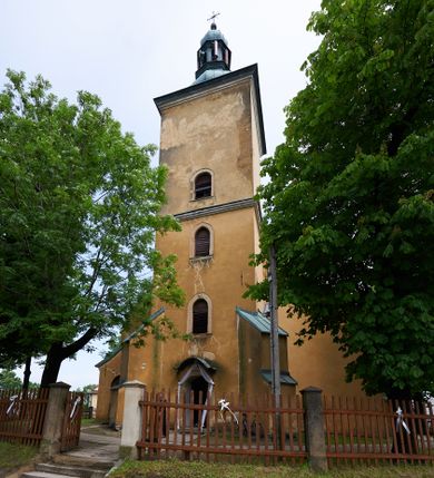 Zdjęcie nr 1: Kościół murowany z cegły i kamienia, tynkowany. Zlokalizowany po północnej stronie wiejskiej drogi, na placu wydzielonym drewnianym płotem rozpiętym pomiędzy murowanymi, tynkowanymi filarami. Budowla orientowana, wzniesiona na wydłużonym rzucie złożonym z krótkiego, zamkniętego wielobocznie prezbiterium, nawy na rzucie zbliżonym  do kwadratu z centralnie umieszczonym filarem i wieży na rzucie kwadratu. Od północy do prezbiterium przylega zakrystia na rzucie kwadratu, do nawy kaplica na rzucie prostokąta. Do wieży dostawiony aneks z klatką schodową prowadzącą na chór. Od południa do nawy przylega kruchta na rzucie kwadratu. Całość budowli oszkarpowana w narożach. Wnętrze nakrywają sklepienia krzyżowe, w nawie żebra wsparte są na czworobocznym, murowanym filarze centralnym. W kruchcie pod wieżą sklepienie kryształowe, w zakrystii sieciowe. Otwory drzwiowe o wykroju ostrołukowym, w kamiennych portalach. Okna o wykroju stojących prostokątów, zwieńczonych łukiem pełnym; w północnej ścianie kaplicy okulus. Elewacje świątyni opracowane jednolicie, tynkowane, z niezbyt wydatnym gzymsem podokapowym, w narożach opięte masywnymi, trójuskokowymi przyporami. Elewacja zachodnia, wieżowa, czterokondygnacyjna, jednoosiowa, flankowana parą przypór. Gzyms kordonowy wydziela ostatnią, najwyższa kondygnację. W osi portal kamienny, zamknięty łukiem półkolistym. Powyżej, w osi okna w każdej z kondygnacji. Na pierwszym piętrze, w zworniku obramienia okiennego herb Drużyna w kartuszu. Boczne elewacje wieży bez otworów okiennych. Elewacja północna artykułowana przyporami, o nieregularnym układzie osi, przesłonięta przybudówkami kaplicy, zakrystii i klatki schodowej wiodącej na chór. W elewacji kaplicy, w osi okulus. W części prezbiterialnej okna o wykroju prostokąta zamkniętego łukiem półkolistym w dekoracyjnych obramieniach kamiennych z pilasterkami i archiwoltą zdobione płycinami i dekoracją ornamentalną w formie rozet i herbem Drużyna. Elewacja wschodnia artykułowana przyporami, przesłonięta parawanową ścianką drewnianą o trzech przeszklonych oknach-arkadah; w głębi ogrójec. Elewacja południowa artykułowana przyporami. W osi korpusu nawowego kruchta, ponad nią, po bokach dwa okna z obramieniami kamiennymi, dekorowanymi ornamentalnie i herbem Drużyna.  Dachy dwuspadowe, kryte blachą. Nieco wyższy nad korpusem nawowym, niższy nad prezbiterium. Nad zakrystią dach pulpitowy. Wieża zwieńczona cebulastym hełmem z latarnią.