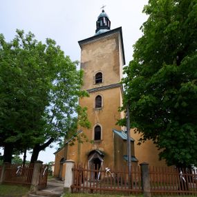 Zdjęcie nr 1: Kościół murowany z cegły i kamienia, tynkowany. Zlokalizowany po północnej stronie wiejskiej drogi, na placu wydzielonym drewnianym płotem rozpiętym pomiędzy murowanymi, tynkowanymi filarami. Budowla orientowana, wzniesiona na wydłużonym rzucie złożonym z krótkiego, zamkniętego wielobocznie prezbiterium, nawy na rzucie zbliżonym  do kwadratu z centralnie umieszczonym filarem i wieży na rzucie kwadratu. Od północy do prezbiterium przylega zakrystia na rzucie kwadratu, do nawy kaplica na rzucie prostokąta. Do wieży dostawiony aneks z klatką schodową prowadzącą na chór. Od południa do nawy przylega kruchta na rzucie kwadratu. Całość budowli oszkarpowana w narożach. Wnętrze nakrywają sklepienia krzyżowe, w nawie żebra wsparte są na czworobocznym, murowanym filarze centralnym. W kruchcie pod wieżą sklepienie kryształowe, w zakrystii sieciowe. Otwory drzwiowe o wykroju ostrołukowym, w kamiennych portalach. Okna o wykroju stojących prostokątów, zwieńczonych łukiem pełnym; w północnej ścianie kaplicy okulus. Elewacje świątyni opracowane jednolicie, tynkowane, z niezbyt wydatnym gzymsem podokapowym, w narożach opięte masywnymi, trójuskokowymi przyporami. Elewacja zachodnia, wieżowa, czterokondygnacyjna, jednoosiowa, flankowana parą przypór. Gzyms kordonowy wydziela ostatnią, najwyższa kondygnację. W osi portal kamienny, zamknięty łukiem półkolistym. Powyżej, w osi okna w każdej z kondygnacji. Na pierwszym piętrze, w zworniku obramienia okiennego herb Drużyna w kartuszu. Boczne elewacje wieży bez otworów okiennych. Elewacja północna artykułowana przyporami, o nieregularnym układzie osi, przesłonięta przybudówkami kaplicy, zakrystii i klatki schodowej wiodącej na chór. W elewacji kaplicy, w osi okulus. W części prezbiterialnej okna o wykroju prostokąta zamkniętego łukiem półkolistym w dekoracyjnych obramieniach kamiennych z pilasterkami i archiwoltą zdobione płycinami i dekoracją ornamentalną w formie rozet i herbem Drużyna. Elewacja wschodnia artykułowana przyporami, przesłonięta parawanową ścianką drewnianą o trzech przeszklonych oknach-arkadah; w głębi ogrójec. Elewacja południowa artykułowana przyporami. W osi korpusu nawowego kruchta, ponad nią, po bokach dwa okna z obramieniami kamiennymi, dekorowanymi ornamentalnie i herbem Drużyna.  Dachy dwuspadowe, kryte blachą. Nieco wyższy nad korpusem nawowym, niższy nad prezbiterium. Nad zakrystią dach pulpitowy. Wieża zwieńczona cebulastym hełmem z latarnią.