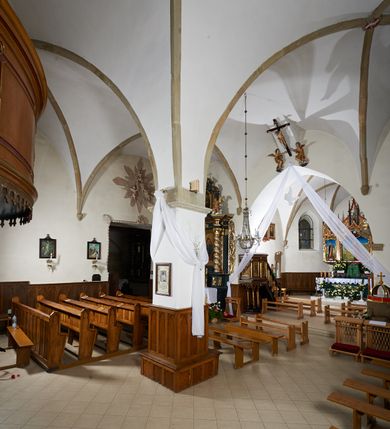 Zdjęcie nr 7: Kościół murowany z cegły i kamienia, tynkowany. Zlokalizowany po północnej stronie wiejskiej drogi, na placu wydzielonym drewnianym płotem rozpiętym pomiędzy murowanymi, tynkowanymi filarami. Budowla orientowana, wzniesiona na wydłużonym rzucie złożonym z krótkiego, zamkniętego wielobocznie prezbiterium, nawy na rzucie zbliżonym  do kwadratu z centralnie umieszczonym filarem i wieży na rzucie kwadratu. Od północy do prezbiterium przylega zakrystia na rzucie kwadratu, do nawy kaplica na rzucie prostokąta. Do wieży dostawiony aneks z klatką schodową prowadzącą na chór. Od południa do nawy przylega kruchta na rzucie kwadratu. Całość budowli oszkarpowana w narożach. Wnętrze nakrywają sklepienia krzyżowe, w nawie żebra wsparte są na czworobocznym, murowanym filarze centralnym. W kruchcie pod wieżą sklepienie kryształowe, w zakrystii sieciowe. Otwory drzwiowe o wykroju ostrołukowym, w kamiennych portalach. Okna o wykroju stojących prostokątów, zwieńczonych łukiem pełnym; w północnej ścianie kaplicy okulus. Elewacje świątyni opracowane jednolicie, tynkowane, z niezbyt wydatnym gzymsem podokapowym, w narożach opięte masywnymi, trójuskokowymi przyporami. Elewacja zachodnia, wieżowa, czterokondygnacyjna, jednoosiowa, flankowana parą przypór. Gzyms kordonowy wydziela ostatnią, najwyższa kondygnację. W osi portal kamienny, zamknięty łukiem półkolistym. Powyżej, w osi okna w każdej z kondygnacji. Na pierwszym piętrze, w zworniku obramienia okiennego herb Drużyna w kartuszu. Boczne elewacje wieży bez otworów okiennych. Elewacja północna artykułowana przyporami, o nieregularnym układzie osi, przesłonięta przybudówkami kaplicy, zakrystii i klatki schodowej wiodącej na chór. W elewacji kaplicy, w osi okulus. W części prezbiterialnej okna o wykroju prostokąta zamkniętego łukiem półkolistym w dekoracyjnych obramieniach kamiennych z pilasterkami i archiwoltą zdobione płycinami i dekoracją ornamentalną w formie rozet i herbem Drużyna. Elewacja wschodnia artykułowana przyporami, przesłonięta parawanową ścianką drewnianą o trzech przeszklonych oknach-arkadah; w głębi ogrójec. Elewacja południowa artykułowana przyporami. W osi korpusu nawowego kruchta, ponad nią, po bokach dwa okna z obramieniami kamiennymi, dekorowanymi ornamentalnie i herbem Drużyna.  Dachy dwuspadowe, kryte blachą. Nieco wyższy nad korpusem nawowym, niższy nad prezbiterium. Nad zakrystią dach pulpitowy. Wieża zwieńczona cebulastym hełmem z latarnią.