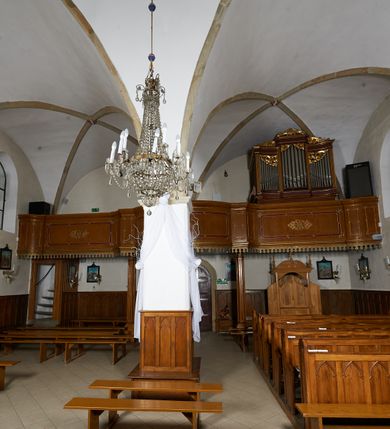 Zdjęcie nr 10: Kościół murowany z cegły i kamienia, tynkowany. Zlokalizowany po północnej stronie wiejskiej drogi, na placu wydzielonym drewnianym płotem rozpiętym pomiędzy murowanymi, tynkowanymi filarami. Budowla orientowana, wzniesiona na wydłużonym rzucie złożonym z krótkiego, zamkniętego wielobocznie prezbiterium, nawy na rzucie zbliżonym  do kwadratu z centralnie umieszczonym filarem i wieży na rzucie kwadratu. Od północy do prezbiterium przylega zakrystia na rzucie kwadratu, do nawy kaplica na rzucie prostokąta. Do wieży dostawiony aneks z klatką schodową prowadzącą na chór. Od południa do nawy przylega kruchta na rzucie kwadratu. Całość budowli oszkarpowana w narożach. Wnętrze nakrywają sklepienia krzyżowe, w nawie żebra wsparte są na czworobocznym, murowanym filarze centralnym. W kruchcie pod wieżą sklepienie kryształowe, w zakrystii sieciowe. Otwory drzwiowe o wykroju ostrołukowym, w kamiennych portalach. Okna o wykroju stojących prostokątów, zwieńczonych łukiem pełnym; w północnej ścianie kaplicy okulus. Elewacje świątyni opracowane jednolicie, tynkowane, z niezbyt wydatnym gzymsem podokapowym, w narożach opięte masywnymi, trójuskokowymi przyporami. Elewacja zachodnia, wieżowa, czterokondygnacyjna, jednoosiowa, flankowana parą przypór. Gzyms kordonowy wydziela ostatnią, najwyższa kondygnację. W osi portal kamienny, zamknięty łukiem półkolistym. Powyżej, w osi okna w każdej z kondygnacji. Na pierwszym piętrze, w zworniku obramienia okiennego herb Drużyna w kartuszu. Boczne elewacje wieży bez otworów okiennych. Elewacja północna artykułowana przyporami, o nieregularnym układzie osi, przesłonięta przybudówkami kaplicy, zakrystii i klatki schodowej wiodącej na chór. W elewacji kaplicy, w osi okulus. W części prezbiterialnej okna o wykroju prostokąta zamkniętego łukiem półkolistym w dekoracyjnych obramieniach kamiennych z pilasterkami i archiwoltą zdobione płycinami i dekoracją ornamentalną w formie rozet i herbem Drużyna. Elewacja wschodnia artykułowana przyporami, przesłonięta parawanową ścianką drewnianą o trzech przeszklonych oknach-arkadah; w głębi ogrójec. Elewacja południowa artykułowana przyporami. W osi korpusu nawowego kruchta, ponad nią, po bokach dwa okna z obramieniami kamiennymi, dekorowanymi ornamentalnie i herbem Drużyna.  Dachy dwuspadowe, kryte blachą. Nieco wyższy nad korpusem nawowym, niższy nad prezbiterium. Nad zakrystią dach pulpitowy. Wieża zwieńczona cebulastym hełmem z latarnią.