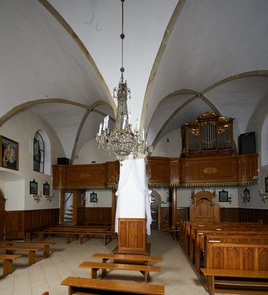 Zdjęcie nr 9: Kościół murowany z cegły i kamienia, tynkowany. Zlokalizowany po północnej stronie wiejskiej drogi, na placu wydzielonym drewnianym płotem rozpiętym pomiędzy murowanymi, tynkowanymi filarami. Budowla orientowana, wzniesiona na wydłużonym rzucie złożonym z krótkiego, zamkniętego wielobocznie prezbiterium, nawy na rzucie zbliżonym  do kwadratu z centralnie umieszczonym filarem i wieży na rzucie kwadratu. Od północy do prezbiterium przylega zakrystia na rzucie kwadratu, do nawy kaplica na rzucie prostokąta. Do wieży dostawiony aneks z klatką schodową prowadzącą na chór. Od południa do nawy przylega kruchta na rzucie kwadratu. Całość budowli oszkarpowana w narożach. Wnętrze nakrywają sklepienia krzyżowe, w nawie żebra wsparte są na czworobocznym, murowanym filarze centralnym. W kruchcie pod wieżą sklepienie kryształowe, w zakrystii sieciowe. Otwory drzwiowe o wykroju ostrołukowym, w kamiennych portalach. Okna o wykroju stojących prostokątów, zwieńczonych łukiem pełnym; w północnej ścianie kaplicy okulus. Elewacje świątyni opracowane jednolicie, tynkowane, z niezbyt wydatnym gzymsem podokapowym, w narożach opięte masywnymi, trójuskokowymi przyporami. Elewacja zachodnia, wieżowa, czterokondygnacyjna, jednoosiowa, flankowana parą przypór. Gzyms kordonowy wydziela ostatnią, najwyższa kondygnację. W osi portal kamienny, zamknięty łukiem półkolistym. Powyżej, w osi okna w każdej z kondygnacji. Na pierwszym piętrze, w zworniku obramienia okiennego herb Drużyna w kartuszu. Boczne elewacje wieży bez otworów okiennych. Elewacja północna artykułowana przyporami, o nieregularnym układzie osi, przesłonięta przybudówkami kaplicy, zakrystii i klatki schodowej wiodącej na chór. W elewacji kaplicy, w osi okulus. W części prezbiterialnej okna o wykroju prostokąta zamkniętego łukiem półkolistym w dekoracyjnych obramieniach kamiennych z pilasterkami i archiwoltą zdobione płycinami i dekoracją ornamentalną w formie rozet i herbem Drużyna. Elewacja wschodnia artykułowana przyporami, przesłonięta parawanową ścianką drewnianą o trzech przeszklonych oknach-arkadah; w głębi ogrójec. Elewacja południowa artykułowana przyporami. W osi korpusu nawowego kruchta, ponad nią, po bokach dwa okna z obramieniami kamiennymi, dekorowanymi ornamentalnie i herbem Drużyna.  Dachy dwuspadowe, kryte blachą. Nieco wyższy nad korpusem nawowym, niższy nad prezbiterium. Nad zakrystią dach pulpitowy. Wieża zwieńczona cebulastym hełmem z latarnią.