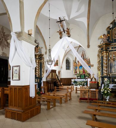 Zdjęcie nr 5: Kościół murowany z cegły i kamienia, tynkowany. Zlokalizowany po północnej stronie wiejskiej drogi, na placu wydzielonym drewnianym płotem rozpiętym pomiędzy murowanymi, tynkowanymi filarami. Budowla orientowana, wzniesiona na wydłużonym rzucie złożonym z krótkiego, zamkniętego wielobocznie prezbiterium, nawy na rzucie zbliżonym  do kwadratu z centralnie umieszczonym filarem i wieży na rzucie kwadratu. Od północy do prezbiterium przylega zakrystia na rzucie kwadratu, do nawy kaplica na rzucie prostokąta. Do wieży dostawiony aneks z klatką schodową prowadzącą na chór. Od południa do nawy przylega kruchta na rzucie kwadratu. Całość budowli oszkarpowana w narożach. Wnętrze nakrywają sklepienia krzyżowe, w nawie żebra wsparte są na czworobocznym, murowanym filarze centralnym. W kruchcie pod wieżą sklepienie kryształowe, w zakrystii sieciowe. Otwory drzwiowe o wykroju ostrołukowym, w kamiennych portalach. Okna o wykroju stojących prostokątów, zwieńczonych łukiem pełnym; w północnej ścianie kaplicy okulus. Elewacje świątyni opracowane jednolicie, tynkowane, z niezbyt wydatnym gzymsem podokapowym, w narożach opięte masywnymi, trójuskokowymi przyporami. Elewacja zachodnia, wieżowa, czterokondygnacyjna, jednoosiowa, flankowana parą przypór. Gzyms kordonowy wydziela ostatnią, najwyższa kondygnację. W osi portal kamienny, zamknięty łukiem półkolistym. Powyżej, w osi okna w każdej z kondygnacji. Na pierwszym piętrze, w zworniku obramienia okiennego herb Drużyna w kartuszu. Boczne elewacje wieży bez otworów okiennych. Elewacja północna artykułowana przyporami, o nieregularnym układzie osi, przesłonięta przybudówkami kaplicy, zakrystii i klatki schodowej wiodącej na chór. W elewacji kaplicy, w osi okulus. W części prezbiterialnej okna o wykroju prostokąta zamkniętego łukiem półkolistym w dekoracyjnych obramieniach kamiennych z pilasterkami i archiwoltą zdobione płycinami i dekoracją ornamentalną w formie rozet i herbem Drużyna. Elewacja wschodnia artykułowana przyporami, przesłonięta parawanową ścianką drewnianą o trzech przeszklonych oknach-arkadah; w głębi ogrójec. Elewacja południowa artykułowana przyporami. W osi korpusu nawowego kruchta, ponad nią, po bokach dwa okna z obramieniami kamiennymi, dekorowanymi ornamentalnie i herbem Drużyna.  Dachy dwuspadowe, kryte blachą. Nieco wyższy nad korpusem nawowym, niższy nad prezbiterium. Nad zakrystią dach pulpitowy. Wieża zwieńczona cebulastym hełmem z latarnią.