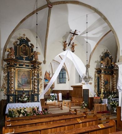 Zdjęcie nr 6: Kościół murowany z cegły i kamienia, tynkowany. Zlokalizowany po północnej stronie wiejskiej drogi, na placu wydzielonym drewnianym płotem rozpiętym pomiędzy murowanymi, tynkowanymi filarami. Budowla orientowana, wzniesiona na wydłużonym rzucie złożonym z krótkiego, zamkniętego wielobocznie prezbiterium, nawy na rzucie zbliżonym  do kwadratu z centralnie umieszczonym filarem i wieży na rzucie kwadratu. Od północy do prezbiterium przylega zakrystia na rzucie kwadratu, do nawy kaplica na rzucie prostokąta. Do wieży dostawiony aneks z klatką schodową prowadzącą na chór. Od południa do nawy przylega kruchta na rzucie kwadratu. Całość budowli oszkarpowana w narożach. Wnętrze nakrywają sklepienia krzyżowe, w nawie żebra wsparte są na czworobocznym, murowanym filarze centralnym. W kruchcie pod wieżą sklepienie kryształowe, w zakrystii sieciowe. Otwory drzwiowe o wykroju ostrołukowym, w kamiennych portalach. Okna o wykroju stojących prostokątów, zwieńczonych łukiem pełnym; w północnej ścianie kaplicy okulus. Elewacje świątyni opracowane jednolicie, tynkowane, z niezbyt wydatnym gzymsem podokapowym, w narożach opięte masywnymi, trójuskokowymi przyporami. Elewacja zachodnia, wieżowa, czterokondygnacyjna, jednoosiowa, flankowana parą przypór. Gzyms kordonowy wydziela ostatnią, najwyższa kondygnację. W osi portal kamienny, zamknięty łukiem półkolistym. Powyżej, w osi okna w każdej z kondygnacji. Na pierwszym piętrze, w zworniku obramienia okiennego herb Drużyna w kartuszu. Boczne elewacje wieży bez otworów okiennych. Elewacja północna artykułowana przyporami, o nieregularnym układzie osi, przesłonięta przybudówkami kaplicy, zakrystii i klatki schodowej wiodącej na chór. W elewacji kaplicy, w osi okulus. W części prezbiterialnej okna o wykroju prostokąta zamkniętego łukiem półkolistym w dekoracyjnych obramieniach kamiennych z pilasterkami i archiwoltą zdobione płycinami i dekoracją ornamentalną w formie rozet i herbem Drużyna. Elewacja wschodnia artykułowana przyporami, przesłonięta parawanową ścianką drewnianą o trzech przeszklonych oknach-arkadah; w głębi ogrójec. Elewacja południowa artykułowana przyporami. W osi korpusu nawowego kruchta, ponad nią, po bokach dwa okna z obramieniami kamiennymi, dekorowanymi ornamentalnie i herbem Drużyna.  Dachy dwuspadowe, kryte blachą. Nieco wyższy nad korpusem nawowym, niższy nad prezbiterium. Nad zakrystią dach pulpitowy. Wieża zwieńczona cebulastym hełmem z latarnią.