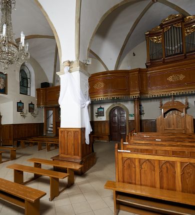 Zdjęcie nr 11: Kościół murowany z cegły i kamienia, tynkowany. Zlokalizowany po północnej stronie wiejskiej drogi, na placu wydzielonym drewnianym płotem rozpiętym pomiędzy murowanymi, tynkowanymi filarami. Budowla orientowana, wzniesiona na wydłużonym rzucie złożonym z krótkiego, zamkniętego wielobocznie prezbiterium, nawy na rzucie zbliżonym  do kwadratu z centralnie umieszczonym filarem i wieży na rzucie kwadratu. Od północy do prezbiterium przylega zakrystia na rzucie kwadratu, do nawy kaplica na rzucie prostokąta. Do wieży dostawiony aneks z klatką schodową prowadzącą na chór. Od południa do nawy przylega kruchta na rzucie kwadratu. Całość budowli oszkarpowana w narożach. Wnętrze nakrywają sklepienia krzyżowe, w nawie żebra wsparte są na czworobocznym, murowanym filarze centralnym. W kruchcie pod wieżą sklepienie kryształowe, w zakrystii sieciowe. Otwory drzwiowe o wykroju ostrołukowym, w kamiennych portalach. Okna o wykroju stojących prostokątów, zwieńczonych łukiem pełnym; w północnej ścianie kaplicy okulus. Elewacje świątyni opracowane jednolicie, tynkowane, z niezbyt wydatnym gzymsem podokapowym, w narożach opięte masywnymi, trójuskokowymi przyporami. Elewacja zachodnia, wieżowa, czterokondygnacyjna, jednoosiowa, flankowana parą przypór. Gzyms kordonowy wydziela ostatnią, najwyższa kondygnację. W osi portal kamienny, zamknięty łukiem półkolistym. Powyżej, w osi okna w każdej z kondygnacji. Na pierwszym piętrze, w zworniku obramienia okiennego herb Drużyna w kartuszu. Boczne elewacje wieży bez otworów okiennych. Elewacja północna artykułowana przyporami, o nieregularnym układzie osi, przesłonięta przybudówkami kaplicy, zakrystii i klatki schodowej wiodącej na chór. W elewacji kaplicy, w osi okulus. W części prezbiterialnej okna o wykroju prostokąta zamkniętego łukiem półkolistym w dekoracyjnych obramieniach kamiennych z pilasterkami i archiwoltą zdobione płycinami i dekoracją ornamentalną w formie rozet i herbem Drużyna. Elewacja wschodnia artykułowana przyporami, przesłonięta parawanową ścianką drewnianą o trzech przeszklonych oknach-arkadah; w głębi ogrójec. Elewacja południowa artykułowana przyporami. W osi korpusu nawowego kruchta, ponad nią, po bokach dwa okna z obramieniami kamiennymi, dekorowanymi ornamentalnie i herbem Drużyna.  Dachy dwuspadowe, kryte blachą. Nieco wyższy nad korpusem nawowym, niższy nad prezbiterium. Nad zakrystią dach pulpitowy. Wieża zwieńczona cebulastym hełmem z latarnią.