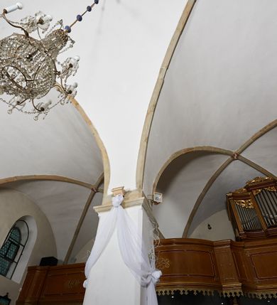 Zdjęcie nr 14: Kościół murowany z cegły i kamienia, tynkowany. Zlokalizowany po północnej stronie wiejskiej drogi, na placu wydzielonym drewnianym płotem rozpiętym pomiędzy murowanymi, tynkowanymi filarami. Budowla orientowana, wzniesiona na wydłużonym rzucie złożonym z krótkiego, zamkniętego wielobocznie prezbiterium, nawy na rzucie zbliżonym  do kwadratu z centralnie umieszczonym filarem i wieży na rzucie kwadratu. Od północy do prezbiterium przylega zakrystia na rzucie kwadratu, do nawy kaplica na rzucie prostokąta. Do wieży dostawiony aneks z klatką schodową prowadzącą na chór. Od południa do nawy przylega kruchta na rzucie kwadratu. Całość budowli oszkarpowana w narożach. Wnętrze nakrywają sklepienia krzyżowe, w nawie żebra wsparte są na czworobocznym, murowanym filarze centralnym. W kruchcie pod wieżą sklepienie kryształowe, w zakrystii sieciowe. Otwory drzwiowe o wykroju ostrołukowym, w kamiennych portalach. Okna o wykroju stojących prostokątów, zwieńczonych łukiem pełnym; w północnej ścianie kaplicy okulus. Elewacje świątyni opracowane jednolicie, tynkowane, z niezbyt wydatnym gzymsem podokapowym, w narożach opięte masywnymi, trójuskokowymi przyporami. Elewacja zachodnia, wieżowa, czterokondygnacyjna, jednoosiowa, flankowana parą przypór. Gzyms kordonowy wydziela ostatnią, najwyższa kondygnację. W osi portal kamienny, zamknięty łukiem półkolistym. Powyżej, w osi okna w każdej z kondygnacji. Na pierwszym piętrze, w zworniku obramienia okiennego herb Drużyna w kartuszu. Boczne elewacje wieży bez otworów okiennych. Elewacja północna artykułowana przyporami, o nieregularnym układzie osi, przesłonięta przybudówkami kaplicy, zakrystii i klatki schodowej wiodącej na chór. W elewacji kaplicy, w osi okulus. W części prezbiterialnej okna o wykroju prostokąta zamkniętego łukiem półkolistym w dekoracyjnych obramieniach kamiennych z pilasterkami i archiwoltą zdobione płycinami i dekoracją ornamentalną w formie rozet i herbem Drużyna. Elewacja wschodnia artykułowana przyporami, przesłonięta parawanową ścianką drewnianą o trzech przeszklonych oknach-arkadah; w głębi ogrójec. Elewacja południowa artykułowana przyporami. W osi korpusu nawowego kruchta, ponad nią, po bokach dwa okna z obramieniami kamiennymi, dekorowanymi ornamentalnie i herbem Drużyna.  Dachy dwuspadowe, kryte blachą. Nieco wyższy nad korpusem nawowym, niższy nad prezbiterium. Nad zakrystią dach pulpitowy. Wieża zwieńczona cebulastym hełmem z latarnią.