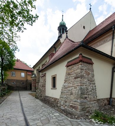 Zdjęcie nr 68: Kościół ceglany, jednonawowy z transeptem. Nawa artykułowana półfilarami i półkolumnami przechodzącymi w gurty sklepienia kolebkowego z lunetami w nawie głównej i żaglowego na przecięciu naw. Na ścianach polichromia na cegle, gurty podkreślone pasami wzorów geometrycznych, sklepienie niebieskie. W nawie i prezbiterium okna w formie biforiów, w transepcie w kształcie stojącego prostokąta zamkniętego łukiem pełnym, z maswerkami, w chórze węższe, zamknięte łukiem ostrym, dwudzielne z rozetą oraz biforia; główne wejście na osi. Chór muzyczny wsparty na masywnych filarach i kolumnach o kapitelach kostkowych z kamiennym parapetem ozdobionym fryzem arkadkowym. Na osi wieloplanowej fasady potężna, trójkondygnacyjna wieża. W pierwszej kondygnacji, po schodach wejście w formie łuku pełnego z drzwiami w formie stojącego prostokąta zamkniętego łukiem dwuramiennym. Obramienie wejścia kamienne, flankowane kolumnami z kostkowymi kapitelami, ze szczytem z chrystogramem. Całość wtopiona w szerszy szczyt, w zwieńczeniu którego rozeta. W trzeciej kondygnacji, zakomponowanej na drugim planie, okno w kształcie stojącego prostokąta zamkniętego łukiem pełnym, dwudzielne z rozetą. Nad nim rzeźba ukrzyżowanego Chrystusa, a w czterech narożach, na konsolach figury św. Stanisława biskupa, św. Stanisława Kostki, św. Jana Kantego i św. Kazimierza Jagiellończyka wykonane z kamienia pińczowskiego przez krakowskiego rzeźbiarza Władysława Druciaka. Wieżę flankują dwie mniejsze, również trójkondygnacyjne wieżyczki dołem czworoboczne, przechodzące w ośmiobok przepruty małymi, prostokątnymi oknami w dwóch niższych kondygnacjach i takimi samymi, naprzemiennie z biforiami w najwyższej. Elewacje transeptu z ogromnymi oknami dekorowanymi maswerkiem, zamknięte trójkątnymi szczytami. Elewacje zewnętrzne z wielobarwnej cegły – w większości ciemnej, dekorowanej na dwóch lub trzech poziomach pasami. Na poziomie pierwszej kondygnacji z jasnej cegły między pasami kamienia, poniżej linii dachu ze wzorem geometrycznym między pasami kamienia, w szczytach jasnym wzorem geometrycznym; chór dekorują blendy arkadkowe. Elewacje opinają ceglane szkarpy, w obejściu zwieńczone kamiennymi kapliczkami. Dachy nawy i transeptu dwuspadowe, dekorowane mozaiką geometryczną, wieloboczna kaplica z dachem sześciokątnym zwieńczonym barokową w kształcie wieżyczką na sygnaturkę, obejście pulpitowym. Do kaplicy przylega przełączka łącząca nowy kościół ze starym.  

Z wyposażenia kościoła na uwagę zasługują obraz Matki Boskiej Suskiej i dwie rzeźby (św. Łukasza i św. Jana Ewangelisty) przeniesione z ołtarza bocznego starego kościoła ustawione na ołtarzu głównym. Sam ołtarz główny (1912) wykonany został przez Antoniego Madeyskiego w Rzymie według rysunków Zygmunta Hendla. Madeyski wykorzystał do wzniesienia ołtarza rzadkie gatunki marmurów z rozebranego starochrześcijańskiego kościoła włoskiego. Niestety przerobiono go i zubożono w 1988 roku. Antoni Madeyski wykonał również tabernakulum, mensę ołtarzową, pulpit oraz chrzcielnicę. Teodor Talowski, architekt, zaprojektował również w 1901 roku ambonę z baldachimem. Zygmunt Hendel zaprojektował, prócz ołtarza głównego, ołtarz boczny św. Józefa i męki Pańskiej (1927). Franciszek Mączyński wykonał figurę Serca Pana Jezusa w latach 1914-1915. Za ołtarzem ze sceną Nawiedzenia widoczne są witraże według kartonów Zdzisława Giedliczki, w prezbiterium ze sceną Zesłania Ducha Świętego i postaciami Matki Boskiej Ostrobramskiej i św. Aleksandra.