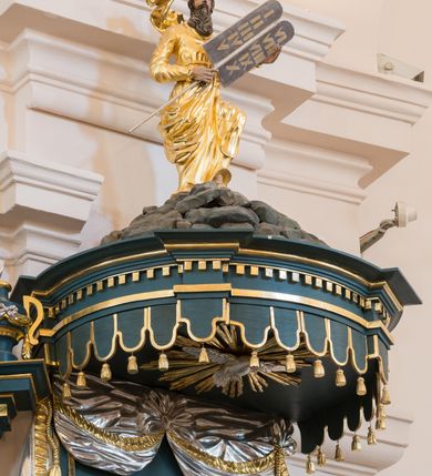 Zdjęcie nr 3: Ambona przyścienna, wisząca. Kosz na planie wycinka koła, dołem wypukły ujęty rzeźbioną formą wieńca laurowego. Trzy ścianki kosza zwężające się ku górze, wydzielone czterema stylizowanymi pilastrami dźwigającymi niepełne belkowanie tworzące parapet kosza, wyłamane na ich osi; trzony dekorowane cekinami, dolna cześć spływająca na cokół tworzy płyciny dekorowane rozetami. Pola zdobione płycinami: dwie zewnętrzne z rozetami przyjmującymi kształt krzyża, środkowa z płaskorzeźbionym przedstawieniem Chrystusa Dobrego pasterza na tle krajobrazu. U spodu kosza podwieszenie w formie spłaszczonej półkuli, podzielonej czterema lizenami na trzy pola dekorowane rozetami, zakończone szyszką. Zaplecek w formie prostokąta ujęty wąskimi lizenami, zwieńczony podwieszonymi płaskorzeźbionymi draperiami, spływającymi fragmentami tkaniny na boki. W polu drzwi ujęte ramą dekorowaną skręconą wstęgą. Baldachim na planie analogicznym do kosza, ujęty na całej długości niepełnym belkowaniem, wyłamanym w czterech miejscach, z podwieszonym lambrekinem. Stanowi on podstawę dla rzeźbionej góry, na której szczycie stoi figura Mojżesza z tablicami przykazań. Na podniebiu baldachimu rzeźbiona gołębica Ducha Świętego w glorii promienistej.
Struktura polichromowana w kolorze ciemnoturkusowym, ornamentyka i profile złocone, detale srebrzone.
