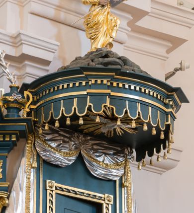 Zdjęcie nr 1: Ambona przyścienna, wisząca. Kosz na planie wycinka koła, dołem wypukły ujęty rzeźbioną formą wieńca laurowego. Trzy ścianki kosza zwężające się ku górze, wydzielone czterema stylizowanymi pilastrami dźwigającymi niepełne belkowanie tworzące parapet kosza, wyłamane na ich osi; trzony dekorowane cekinami, dolna cześć spływająca na cokół tworzy płyciny dekorowane rozetami. Pola zdobione płycinami: dwie zewnętrzne z rozetami przyjmującymi kształt krzyża, środkowa z płaskorzeźbionym przedstawieniem Chrystusa Dobrego pasterza na tle krajobrazu. U spodu kosza podwieszenie w formie spłaszczonej półkuli, podzielonej czterema lizenami na trzy pola dekorowane rozetami, zakończone szyszką. Zaplecek w formie prostokąta ujęty wąskimi lizenami, zwieńczony podwieszonymi płaskorzeźbionymi draperiami, spływającymi fragmentami tkaniny na boki. W polu drzwi ujęte ramą dekorowaną skręconą wstęgą. Baldachim na planie analogicznym do kosza, ujęty na całej długości niepełnym belkowaniem, wyłamanym w czterech miejscach, z podwieszonym lambrekinem. Stanowi on podstawę dla rzeźbionej góry, na której szczycie stoi figura Mojżesza z tablicami przykazań. Na podniebiu baldachimu rzeźbiona gołębica Ducha Świętego w glorii promienistej.
Struktura polichromowana w kolorze ciemnoturkusowym, ornamentyka i profile złocone, detale srebrzone.
