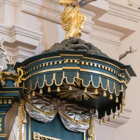 Zdjęcie nr 1: Ambona przyścienna, wisząca. Kosz na planie wycinka koła, dołem wypukły ujęty rzeźbioną formą wieńca laurowego. Trzy ścianki kosza zwężające się ku górze, wydzielone czterema stylizowanymi pilastrami dźwigającymi niepełne belkowanie tworzące parapet kosza, wyłamane na ich osi; trzony dekorowane cekinami, dolna cześć spływająca na cokół tworzy płyciny dekorowane rozetami. Pola zdobione płycinami: dwie zewnętrzne z rozetami przyjmującymi kształt krzyża, środkowa z płaskorzeźbionym przedstawieniem Chrystusa Dobrego pasterza na tle krajobrazu. U spodu kosza podwieszenie w formie spłaszczonej półkuli, podzielonej czterema lizenami na trzy pola dekorowane rozetami, zakończone szyszką. Zaplecek w formie prostokąta ujęty wąskimi lizenami, zwieńczony podwieszonymi płaskorzeźbionymi draperiami, spływającymi fragmentami tkaniny na boki. W polu drzwi ujęte ramą dekorowaną skręconą wstęgą. Baldachim na planie analogicznym do kosza, ujęty na całej długości niepełnym belkowaniem, wyłamanym w czterech miejscach, z podwieszonym lambrekinem. Stanowi on podstawę dla rzeźbionej góry, na której szczycie stoi figura Mojżesza z tablicami przykazań. Na podniebiu baldachimu rzeźbiona gołębica Ducha Świętego w glorii promienistej.
Struktura polichromowana w kolorze ciemnoturkusowym, ornamentyka i profile złocone, detale srebrzone.
