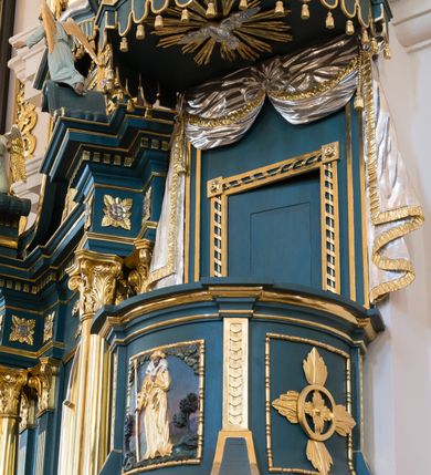 Zdjęcie nr 2: Ambona przyścienna, wisząca. Kosz na planie wycinka koła, dołem wypukły ujęty rzeźbioną formą wieńca laurowego. Trzy ścianki kosza zwężające się ku górze, wydzielone czterema stylizowanymi pilastrami dźwigającymi niepełne belkowanie tworzące parapet kosza, wyłamane na ich osi; trzony dekorowane cekinami, dolna cześć spływająca na cokół tworzy płyciny dekorowane rozetami. Pola zdobione płycinami: dwie zewnętrzne z rozetami przyjmującymi kształt krzyża, środkowa z płaskorzeźbionym przedstawieniem Chrystusa Dobrego pasterza na tle krajobrazu. U spodu kosza podwieszenie w formie spłaszczonej półkuli, podzielonej czterema lizenami na trzy pola dekorowane rozetami, zakończone szyszką. Zaplecek w formie prostokąta ujęty wąskimi lizenami, zwieńczony podwieszonymi płaskorzeźbionymi draperiami, spływającymi fragmentami tkaniny na boki. W polu drzwi ujęte ramą dekorowaną skręconą wstęgą. Baldachim na planie analogicznym do kosza, ujęty na całej długości niepełnym belkowaniem, wyłamanym w czterech miejscach, z podwieszonym lambrekinem. Stanowi on podstawę dla rzeźbionej góry, na której szczycie stoi figura Mojżesza z tablicami przykazań. Na podniebiu baldachimu rzeźbiona gołębica Ducha Świętego w glorii promienistej.
Struktura polichromowana w kolorze ciemnoturkusowym, ornamentyka i profile złocone, detale srebrzone.
