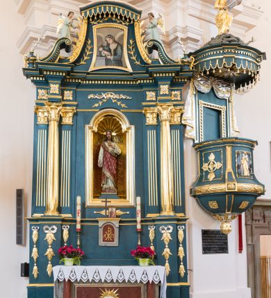 Zdjęcie nr 1: Ołtarz architektoniczny, jednokondygnacyjny, jednoosiowy, ze zwieńczeniem. Ołtarz prostopadłościenny, z drewnianą mensą i kamiennym portatylem. Antenpedium z płyciną w kształcie leżącego prostokąta, ujętą profilowaną, srebrzona ramą, z płaskorzeźbionym, złoconym i srebrzonym przedstawieniem Baranka Paschalnego na tle tarczy słonecznej. Retabulum na wysokim, zdwojonym cokole, o zryzalitowanych, uskokowych bokach, ujęte dwoma parami pilastrów i parą kolumn o kapitelach korynckich i żłobkowanych trzonach, podtrzymujących wyłamujące się ponad nimi, przerwane w partii architrawu i fryzu belkowanie z silnie wysuniętym gzymsem. Zwieńczenie ujęte parą spływów, zamknięte gzymsem w formie nadwieszonego łuku wycinkowego, flankowane fragmentami przerwanego przyczółka. Szczyt w formie promienistej glorii z motywem krzyża pośrodku. Górny cokół retabulum zdobiony motywem przewiązanych wstęgą kampanulli, pośrodku konsolka z polem w kształcie stojącego prostokąta zamkniętego półkoliście z monogramem chrystologicznym (XP AΩ). U góry środkowej części retabulum ornamentyka roślinna z motywem rocaille'u. Partie fryzu zdobione motywem rozet, w partii gzymsu ornament kostkowy. Zwieńczenie dekorowane motywem gałązek liściastych, krawędzie spływów ornamentem cekinowym, w górnej partii lambrekin. Całość flankuje para wazonów. Pole środkowe retabulum w formie prostokątnej niszy zamkniętej konchą o wykroju łuku półkolistego nadwieszonego, ujętej profilowaną, złoconą i srebrzoną ramą. Ścianki niszy zdobione grawerowaną dekoracją stylizowanych motywów roślinnych. Wewnątrz rzeźba Najświętszego Serce Jezusa. W zwieńczeniu pole w kształcie stojącego trapezu z wklęsło-wypukłymi bokami, ujęte profilowaną, złoconą i srebrzoną ramą. Wewnątrz obraz ze św. Rozalią. Na fragmentach przerwanego przyczółka para klęczących aniołków.
 Struktura polichromowana w kolorze ciemnego turkusu. Ołtarz i konsolka retabulum marmoryzowane z odcieniach szarości i brązów. Detal architektoniczny i ornamentyka złocone i srebrzone.     
