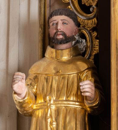 Zdjęcie nr 1: Całopostaciowa, drążona z tyłu figura nieznanego świętego zwróconego na wprost i stojącego w kontrapoście na niewysokiej, prostopadłościennej podstawie. Szeroką twarz charakteryzują delikatne rysy o długim, prostym nosie, małych, szeroko rozstawionych oczach i krótkim zaroście. Włosy są krótkie, ciemnobrązowe. Święty ubrany jest w złocony, drobno fałdowany habit zakonny przewiązany sznurem, na stopach ma brązowe buty. Zgięte w łokciach ręce wyciąga przed siebie.  