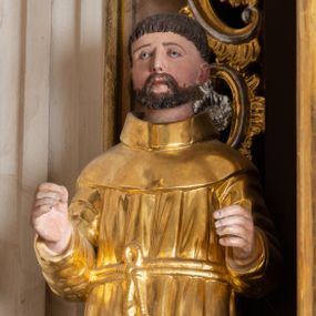 Zdjęcie nr 1: Całopostaciowa, drążona z tyłu figura nieznanego świętego zwróconego na wprost i stojącego w kontrapoście na niewysokiej, prostopadłościennej podstawie. Szeroką twarz charakteryzują delikatne rysy o długim, prostym nosie, małych, szeroko rozstawionych oczach i krótkim zaroście. Włosy są krótkie, ciemnobrązowe. Święty ubrany jest w złocony, drobno fałdowany habit zakonny przewiązany sznurem, na stopach ma brązowe buty. Zgięte w łokciach ręce wyciąga przed siebie.  