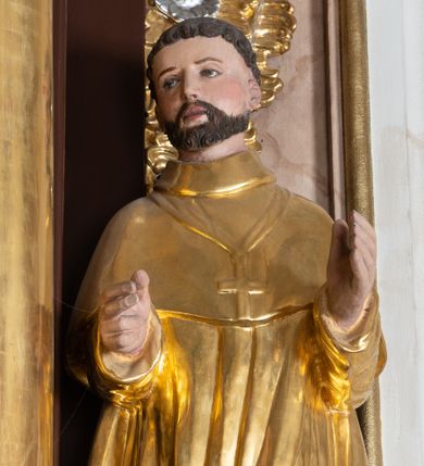 Zdjęcie nr 1: Całopostaciowa, drążona z tyłu figura nieznanego świętego zwróconego na wprost i stojącego w wydatnym kontrapoście na niewysokiej, prostopadłościennej podstawie. Zwróconą w trzech czwartych w prawo głowę charakteryzuje szeroka twarz o delikatnych rysach z długim, prostym nosem i krótkim zarostem. Krótkie, ciemnobrązowe włosy układają się w skręcone pukle. Święty ubrany jest w złocony, drobno fałdowany habit zakonny z peleryną, na piersiach ma złocony krzyż, na stopach brązowe buty. Zgięte w łokciach ręce wyciąga przed siebie.  

