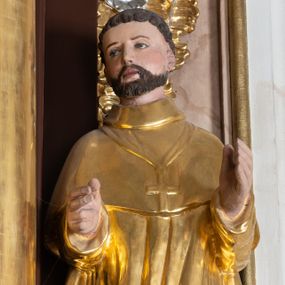 Zdjęcie nr 1: Całopostaciowa, drążona z tyłu figura nieznanego świętego zwróconego na wprost i stojącego w wydatnym kontrapoście na niewysokiej, prostopadłościennej podstawie. Zwróconą w trzech czwartych w prawo głowę charakteryzuje szeroka twarz o delikatnych rysach z długim, prostym nosem i krótkim zarostem. Krótkie, ciemnobrązowe włosy układają się w skręcone pukle. Święty ubrany jest w złocony, drobno fałdowany habit zakonny z peleryną, na piersiach ma złocony krzyż, na stopach brązowe buty. Zgięte w łokciach ręce wyciąga przed siebie.  

