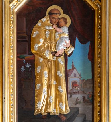 Zdjęcie nr 1: Obraz w kształcie stojącego prostokąta zamkniętego trójbocznie z sześcioboku, ujęty rzeźbioną, złocono-srebrzoną ramą z motywem astragalu, przedstawia św. Antoniego Padewskiego ukazanego całopostaciowo, w trzech czwartych zwróconego w lewo. Pochyloną głowę charakteryzuje owalna twarz o młodzieńczych, delikatnych rysach i opuszczonym wzroku, głowę okala tonsura. Święty trzyma na rękach przytulające się do niego Dzieciątko ujmujące go za podbródek. Jezus zwrócony jest profilem w prawo. Zaokrągloną twarz charakteryzują dziecięce rysy, włosy są krótkie, kręcone. Partie ubiorów postaci oraz ręki i nóg Jezusa oraz lewej dłoni św. Antoniego przesłania metalowa okładzina. W części habitu świętego jest ona złocona i zdobiona srebrzonymi aplikacjami w formie kwiatów, długa suknia Dzieciątka jest srebrzona i przewiązana złoconym paskiem, partie ciała są naturalistycznie polichromowane. Święty Antoni stoi na kamiennym podeście o trzech stopniach, obok niego, po lewej stronie kompozycji znajduje się wysoki wazon z białą lilią. Dalej jest nakryty czerwoną tkaniną stół, na którym leży zamknięta księga w zielonkawej oprawie. W głębi widoczna jest fragmentarycznie kolumna na wysokim cokole oraz ciemnoczerwona kotara podpięta z prawej strony kompozycji. Za nią rozpościera się pejzaż miejski z usytuowanym pośrodku rynku murowanym kościołem z którego wychodzi kapłan pod baldachimem. Ukazane na rynku postaci i zwierzęta przyklękają. W lewym dolnym rogu obrazu sygnatura artysty „T(eodor) Pitala / 26-5-(1)936” .