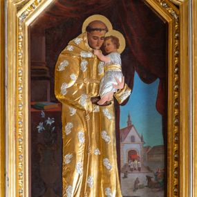 Zdjęcie nr 1: Obraz w kształcie stojącego prostokąta zamkniętego trójbocznie z sześcioboku, ujęty rzeźbioną, złocono-srebrzoną ramą z motywem astragalu, przedstawia św. Antoniego Padewskiego ukazanego całopostaciowo, w trzech czwartych zwróconego w lewo. Pochyloną głowę charakteryzuje owalna twarz o młodzieńczych, delikatnych rysach i opuszczonym wzroku, głowę okala tonsura. Święty trzyma na rękach przytulające się do niego Dzieciątko ujmujące go za podbródek. Jezus zwrócony jest profilem w prawo. Zaokrągloną twarz charakteryzują dziecięce rysy, włosy są krótkie, kręcone. Partie ubiorów postaci oraz ręki i nóg Jezusa oraz lewej dłoni św. Antoniego przesłania metalowa okładzina. W części habitu świętego jest ona złocona i zdobiona srebrzonymi aplikacjami w formie kwiatów, długa suknia Dzieciątka jest srebrzona i przewiązana złoconym paskiem, partie ciała są naturalistycznie polichromowane. Święty Antoni stoi na kamiennym podeście o trzech stopniach, obok niego, po lewej stronie kompozycji znajduje się wysoki wazon z białą lilią. Dalej jest nakryty czerwoną tkaniną stół, na którym leży zamknięta księga w zielonkawej oprawie. W głębi widoczna jest fragmentarycznie kolumna na wysokim cokole oraz ciemnoczerwona kotara podpięta z prawej strony kompozycji. Za nią rozpościera się pejzaż miejski z usytuowanym pośrodku rynku murowanym kościołem z którego wychodzi kapłan pod baldachimem. Ukazane na rynku postaci i zwierzęta przyklękają. W lewym dolnym rogu obrazu sygnatura artysty „T(eodor) Pitala / 26-5-(1)936” .