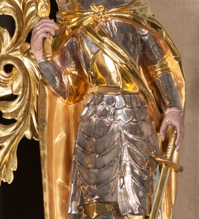 Zdjęcie nr 2: Całopostaciowa, ścięta z tyłu figura św. Wacława stojącego na sferycznej podstawie i zwróconego na wprost. Sylwetka świętego jest w S-kształtnym układzie. Pociągłą twarz charakteryzują wyraziste rysy o małych oczach, długim nosie, zaróżowionych policzkach i krótkim, ciemnym zaroście. Ciemnobrązowe włosy skręcone w pukle rytmicznie opadające sięgając ramion. Święty ubrany jest w srebrzoną zbroję, przez prawe ramię przewiązana diagonalnie złota wstęga, spięty na piersiach, złocony płaszcz opada na plecy i ramiona. Na głowie złocona mitra książęca. W prawej, zgiętej w łokciu i uniesionej do góry ręce trzyma złocone berło, w lewej, opuszczonej wzdłuż ciała złocony miecz.    