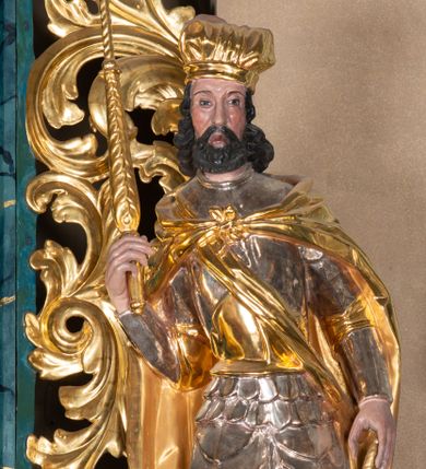 Zdjęcie nr 1: Całopostaciowa, ścięta z tyłu figura św. Wacława stojącego na sferycznej podstawie i zwróconego na wprost. Sylwetka świętego jest w S-kształtnym układzie. Pociągłą twarz charakteryzują wyraziste rysy o małych oczach, długim nosie, zaróżowionych policzkach i krótkim, ciemnym zaroście. Ciemnobrązowe włosy skręcone w pukle rytmicznie opadające sięgając ramion. Święty ubrany jest w srebrzoną zbroję, przez prawe ramię przewiązana diagonalnie złota wstęga, spięty na piersiach, złocony płaszcz opada na plecy i ramiona. Na głowie złocona mitra książęca. W prawej, zgiętej w łokciu i uniesionej do góry ręce trzyma złocone berło, w lewej, opuszczonej wzdłuż ciała złocony miecz.    