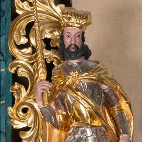 Zdjęcie nr 1: Całopostaciowa, ścięta z tyłu figura św. Wacława stojącego na sferycznej podstawie i zwróconego na wprost. Sylwetka świętego jest w S-kształtnym układzie. Pociągłą twarz charakteryzują wyraziste rysy o małych oczach, długim nosie, zaróżowionych policzkach i krótkim, ciemnym zaroście. Ciemnobrązowe włosy skręcone w pukle rytmicznie opadające sięgając ramion. Święty ubrany jest w srebrzoną zbroję, przez prawe ramię przewiązana diagonalnie złota wstęga, spięty na piersiach, złocony płaszcz opada na plecy i ramiona. Na głowie złocona mitra książęca. W prawej, zgiętej w łokciu i uniesionej do góry ręce trzyma złocone berło, w lewej, opuszczonej wzdłuż ciała złocony miecz.    