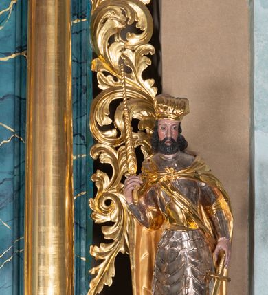 Zdjęcie nr 5: Całopostaciowa, ścięta z tyłu figura św. Wacława stojącego na sferycznej podstawie i zwróconego na wprost. Sylwetka świętego jest w S-kształtnym układzie. Pociągłą twarz charakteryzują wyraziste rysy o małych oczach, długim nosie, zaróżowionych policzkach i krótkim, ciemnym zaroście. Ciemnobrązowe włosy skręcone w pukle rytmicznie opadające sięgając ramion. Święty ubrany jest w srebrzoną zbroję, przez prawe ramię przewiązana diagonalnie złota wstęga, spięty na piersiach, złocony płaszcz opada na plecy i ramiona. Na głowie złocona mitra książęca. W prawej, zgiętej w łokciu i uniesionej do góry ręce trzyma złocone berło, w lewej, opuszczonej wzdłuż ciała złocony miecz.    