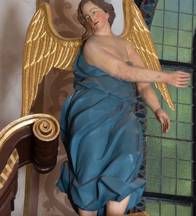 Zdjęcie nr 7: Ołtarz architektoniczny, jednokondygnacyjny, jednoosiowy ze zwieńczeniem i z parą bramek. Ołtarz murowany, prostopadłościenny z malowanym na płótnie antepedium ujętym drewnianą, profilowaną ramą. Wewnątrz przedstawienie wkomponowane w trójprzelotową arkadę z wizerunkiem Jezusa pośrodku i sceną Zwiastowania po bokach. Osadzone na niewysokim cokole tabernakulum jest w formie prostopadłościennej szafki o wypukłej ściance frontowej i narożach artykułowanych skośnie ustawionymi pilastrami wspierającymi wyłamujący się ponad nimi gzyms wieńczący przybierający po środku formę stykających się wolut z motywem konchy. Kwadratowe drzwiczki zamknięte są półkolistym łukiem nadwieszonym. Tabernakulum flankuje para wydatnych spływów wolutowych o krawędziach zdobionych motywem rocaille’owego grzebienia. Struktura jest srebrzona i złocona. Retabulum osadzone na zdwojonym, ukośnie zryzalitowanym cokole z otworami bramek na bokach. Część środkową nastawy ujmują dwie pary kolumn o kapitelach korynckich ustawione na tle pary filarów o kanelowanych trzonach, całość wspiera wyłamujące się ponad nimi przerwane w partii architrawu i fryzu belkowanie z silnie wysuniętym gzymsem. Zwieńczenie w formie przerwanego  przyczółka w kształcie S-kształtnych wolut. Górną partię cokołu zdobią motywy rocaille’ów, natomiast fryz motywy rozet. Pośrodku zwieńczenia usytuowana jest okrągła, promienista gloria z monogramem imienia Jezus i gorejącym sercem pośrodku. Po bokach zwieńczenia stoi para figur aniołów ubranych w niebieskie szaty. Pole środkowe retabulum w formie wnęki w kształcie stojącego prostokąta zamkniętego łukiem koszowym. Wewnątrz krucyfiks na polichromowanym tle przestawiającym widok miasta z murami warownymi i wydatną bryłą kościoła po prawej stronie kompozycji, pokrytym skłębionymi chmurami niebem oraz tarczami - słoneczną po prawej i księżycową po lewej stronie górnej partii krucyfiksu. Struktura ołtarza  jest w kolorze brązowym, detal architektoniczny i ornamentyka są złocone i srebrzone.      