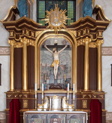 Zdjęcie nr 2: Ołtarz architektoniczny, jednokondygnacyjny, jednoosiowy ze zwieńczeniem i z parą bramek. Ołtarz murowany, prostopadłościenny z malowanym na płótnie antepedium ujętym drewnianą, profilowaną ramą. Wewnątrz przedstawienie wkomponowane w trójprzelotową arkadę z wizerunkiem Jezusa pośrodku i sceną Zwiastowania po bokach. Osadzone na niewysokim cokole tabernakulum jest w formie prostopadłościennej szafki o wypukłej ściance frontowej i narożach artykułowanych skośnie ustawionymi pilastrami wspierającymi wyłamujący się ponad nimi gzyms wieńczący przybierający po środku formę stykających się wolut z motywem konchy. Kwadratowe drzwiczki zamknięte są półkolistym łukiem nadwieszonym. Tabernakulum flankuje para wydatnych spływów wolutowych o krawędziach zdobionych motywem rocaille’owego grzebienia. Struktura jest srebrzona i złocona. Retabulum osadzone na zdwojonym, ukośnie zryzalitowanym cokole z otworami bramek na bokach. Część środkową nastawy ujmują dwie pary kolumn o kapitelach korynckich ustawione na tle pary filarów o kanelowanych trzonach, całość wspiera wyłamujące się ponad nimi przerwane w partii architrawu i fryzu belkowanie z silnie wysuniętym gzymsem. Zwieńczenie w formie przerwanego  przyczółka w kształcie S-kształtnych wolut. Górną partię cokołu zdobią motywy rocaille’ów, natomiast fryz motywy rozet. Pośrodku zwieńczenia usytuowana jest okrągła, promienista gloria z monogramem imienia Jezus i gorejącym sercem pośrodku. Po bokach zwieńczenia stoi para figur aniołów ubranych w niebieskie szaty. Pole środkowe retabulum w formie wnęki w kształcie stojącego prostokąta zamkniętego łukiem koszowym. Wewnątrz krucyfiks na polichromowanym tle przestawiającym widok miasta z murami warownymi i wydatną bryłą kościoła po prawej stronie kompozycji, pokrytym skłębionymi chmurami niebem oraz tarczami - słoneczną po prawej i księżycową po lewej stronie górnej partii krucyfiksu. Struktura ołtarza  jest w kolorze brązowym, detal architektoniczny i ornamentyka są złocone i srebrzone.      