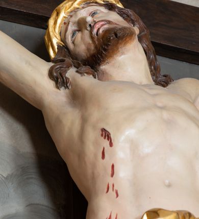 Zdjęcie nr 4: Ołtarz architektoniczny, jednokondygnacyjny, jednoosiowy ze zwieńczeniem i z parą bramek. Ołtarz murowany, prostopadłościenny z malowanym na płótnie antepedium ujętym drewnianą, profilowaną ramą. Wewnątrz przedstawienie wkomponowane w trójprzelotową arkadę z wizerunkiem Jezusa pośrodku i sceną Zwiastowania po bokach. Osadzone na niewysokim cokole tabernakulum jest w formie prostopadłościennej szafki o wypukłej ściance frontowej i narożach artykułowanych skośnie ustawionymi pilastrami wspierającymi wyłamujący się ponad nimi gzyms wieńczący przybierający po środku formę stykających się wolut z motywem konchy. Kwadratowe drzwiczki zamknięte są półkolistym łukiem nadwieszonym. Tabernakulum flankuje para wydatnych spływów wolutowych o krawędziach zdobionych motywem rocaille’owego grzebienia. Struktura jest srebrzona i złocona. Retabulum osadzone na zdwojonym, ukośnie zryzalitowanym cokole z otworami bramek na bokach. Część środkową nastawy ujmują dwie pary kolumn o kapitelach korynckich ustawione na tle pary filarów o kanelowanych trzonach, całość wspiera wyłamujące się ponad nimi przerwane w partii architrawu i fryzu belkowanie z silnie wysuniętym gzymsem. Zwieńczenie w formie przerwanego  przyczółka w kształcie S-kształtnych wolut. Górną partię cokołu zdobią motywy rocaille’ów, natomiast fryz motywy rozet. Pośrodku zwieńczenia usytuowana jest okrągła, promienista gloria z monogramem imienia Jezus i gorejącym sercem pośrodku. Po bokach zwieńczenia stoi para figur aniołów ubranych w niebieskie szaty. Pole środkowe retabulum w formie wnęki w kształcie stojącego prostokąta zamkniętego łukiem koszowym. Wewnątrz krucyfiks na polichromowanym tle przestawiającym widok miasta z murami warownymi i wydatną bryłą kościoła po prawej stronie kompozycji, pokrytym skłębionymi chmurami niebem oraz tarczami - słoneczną po prawej i księżycową po lewej stronie górnej partii krucyfiksu. Struktura ołtarza  jest w kolorze brązowym, detal architektoniczny i ornamentyka są złocone i srebrzone.      