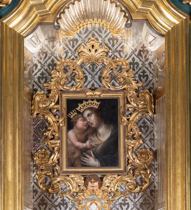 Zdjęcie nr 2: Obraz w kształcie stojącego prostokąta, ujęty profilowaną i ażurową, akantową, złoconą ramą, z przedstawieniem Matki Boskiej z Dzieciątkiem. Maria ukazana w półpostaci, w trzech czwartych zwrócona i lekko pochylona w prawo. Głowa w trzech czwartych przechylona w prawo. Twarz owalna o delikatnych, pełnych rysach. Włosy długie, ciemnobrązowe. Maria ubrana w białą tunikę i otulona ciemnoniebieskim płaszczem. Lewą ręką, podniesiona na wysokości klatki piersiowej, wskazuje na stojące po jej prawej stronie Dzieciątko. Jezus w trzech czwartych zwrócony w lewo, głowę o zaokrąglonej twarzy i z krótkimi, blond, kręconymi włosami, przytula do twarzy Marii. Lewą rączką obejmuje szyję Marii, prawą trzyma połę jej płaszcza. Ciało Dzieciątka od pasa przesłonięte delikatną, przejrzystą tkaniną. Tło ciemne. Na głowach postaci nakładane złocone korony. 
Kolorystyka wysublimowana, stonowana, niezbyt nasyconej tonacji.   
